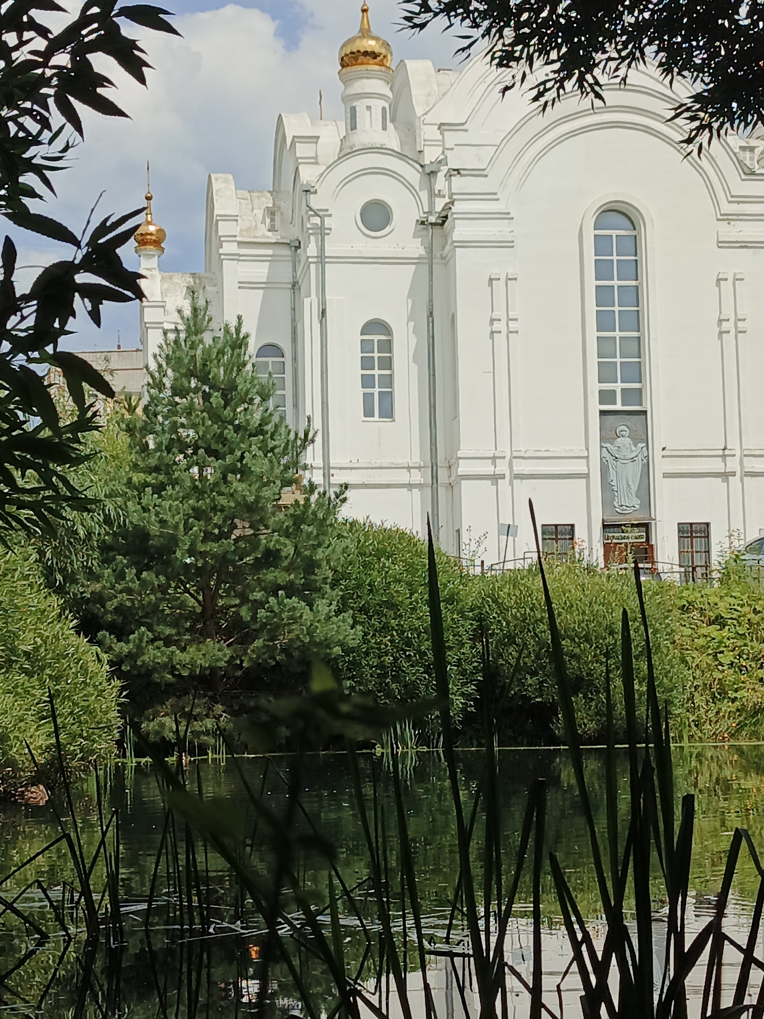 The image depicts the exterior of a white Orthodox church with gold domes, likely located in Zlatoust, Russia. In the foreground, a tranquil pond or body of water is visible, with tall reeds and aquatic plants silhouetted against its surface. The water reflects the surrounding lush green foliage and the bright sky. Behind the water, a dense green hedge borders the church grounds, and a tall evergreen tree stands prominently in front of the building. The church itself is a multi-story structure with arched windows and decorative architectural elements. A relief image of a robed figure is mounted on the facade above an entrance. Dark green foliage from trees in the foreground partially obscures the view of the sky and the upper parts of the church. The scene suggests a serene and picturesque setting, possibly a park or garden adjacent to the religious building.