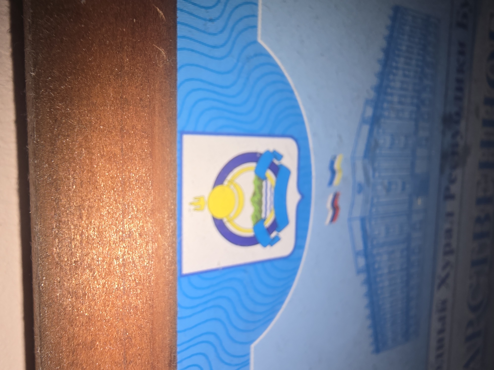 A close-up, slightly angled shot displays a portion of a document or official banner with blue wave-like patterns. In the center, a coat of arms is visible, featuring a yellow sun behind a green landscape with water, all enclosed within a blue and yellow ring. Below the coat of arms, a blue silhouette of a building with numerous windows is depicted, with two flags flying above it: one Russian tricolor and one blue and yellow flag. Further text in a dark color is present at the bottom of the frame, partially obscured. The upper portion of the image shows a textured brown surface, likely wood or a similar material. Based on the coat of arms and flags, the document likely pertains to Ulan-Ude, the capital city of Buryatia, Russia.
