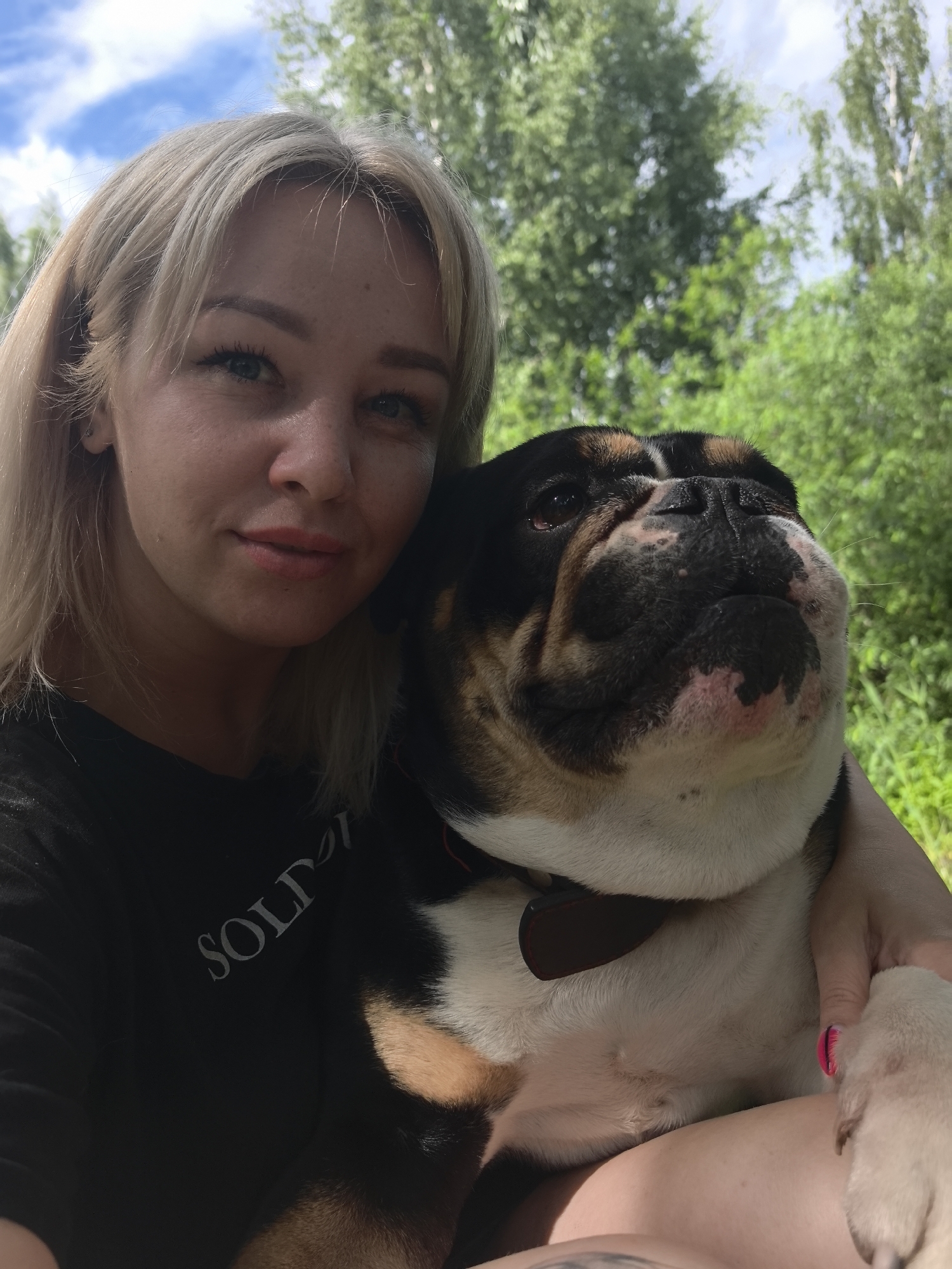 A blonde woman, wearing a black t-shirt with a white graphic reading "SOLD," is holding an English bulldog in her arms. She is looking towards the camera with a slight smile. The dog is looking up and to the right, with its tongue slightly out. They are outdoors, with lush green trees and a blue sky with white clouds in the background. The lighting suggests it is daytime. The specific location of Noviy, Russia, cannot be determined from the image.