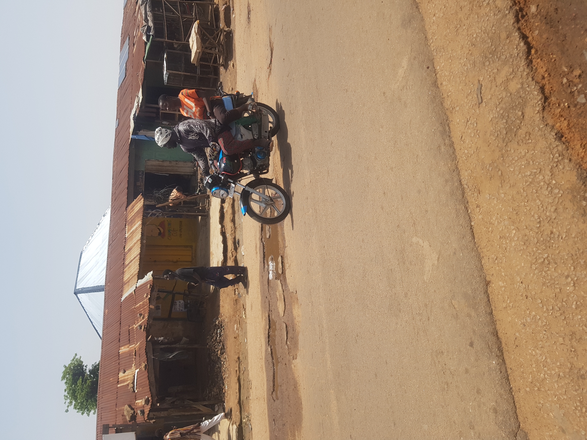 In Tukur Tukur, Nigeria, a motorcycle with two occupants travels on a paved road. The driver, a man wearing a dark patterned shirt and a white headscarf, operates the motorcycle. Behind him, a passenger, a woman wearing an orange t-shirt, sits on the back. The road is in poor condition, with several large potholes filled with murky water. In the background, a row of buildings with corrugated iron roofs and weathered walls lines the street. Some of the buildings appear to be shops, with open fronts displaying various goods or services. A man in a striped shirt and white pants walks on the far left of the image, and another man in dark clothing walks on the right. The sky is clear and the sun is bright.