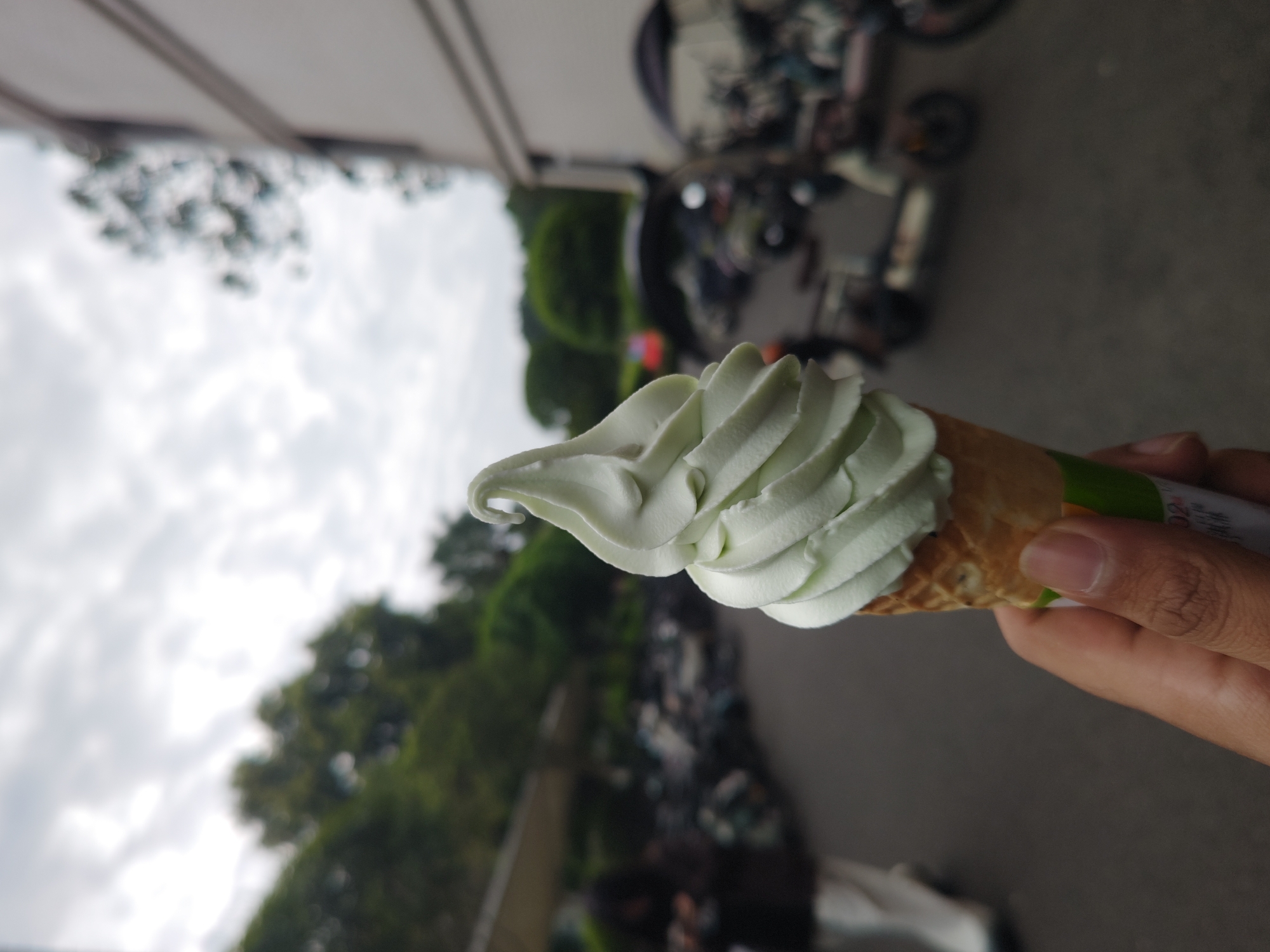 A person is holding a soft-serve ice cream cone. The ice cream is pale green and swirled into a peak. The cone is a waffle cone. The person's hand and part of their arm are visible. The background is blurred, showing trees, a building, and parked motorbikes. The image suggests an outdoor setting in a city.
The photograph appears to be taken in Chongqing High-tech Zone, China.
