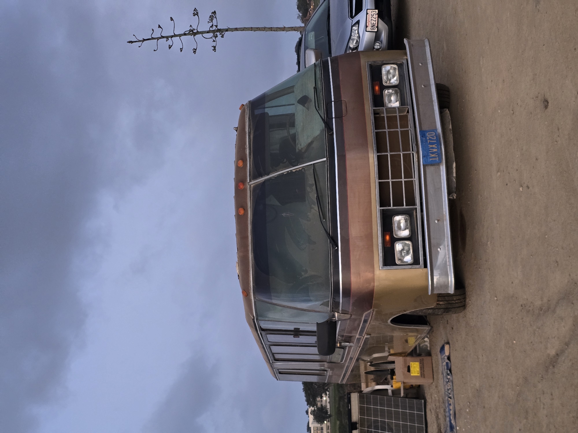 A wide shot captures a vintage, two-tone brown and gold RV parked on a dirt lot under a cloudy, overcast sky. The RV, with its distinctive chrome grille and large panoramic windshield, dominates the foreground. To its left, several solar panels are propped up. In the background, a silver sedan with a license plate reading "8NEZ253" is parked to the right of the RV. Further behind, a tall, bare agave plant stands silhouetted against the sky, and a distant building with trees is visible. The license plate on the RV reads "1KVX720". The overall scene suggests an outdoor, possibly rural or undeveloped area, consistent with a location on the outskirts of San Diego, California, United States of America, perhaps an RV park or a private property where alternative living arrangements are possible. There are no people visible in the image.