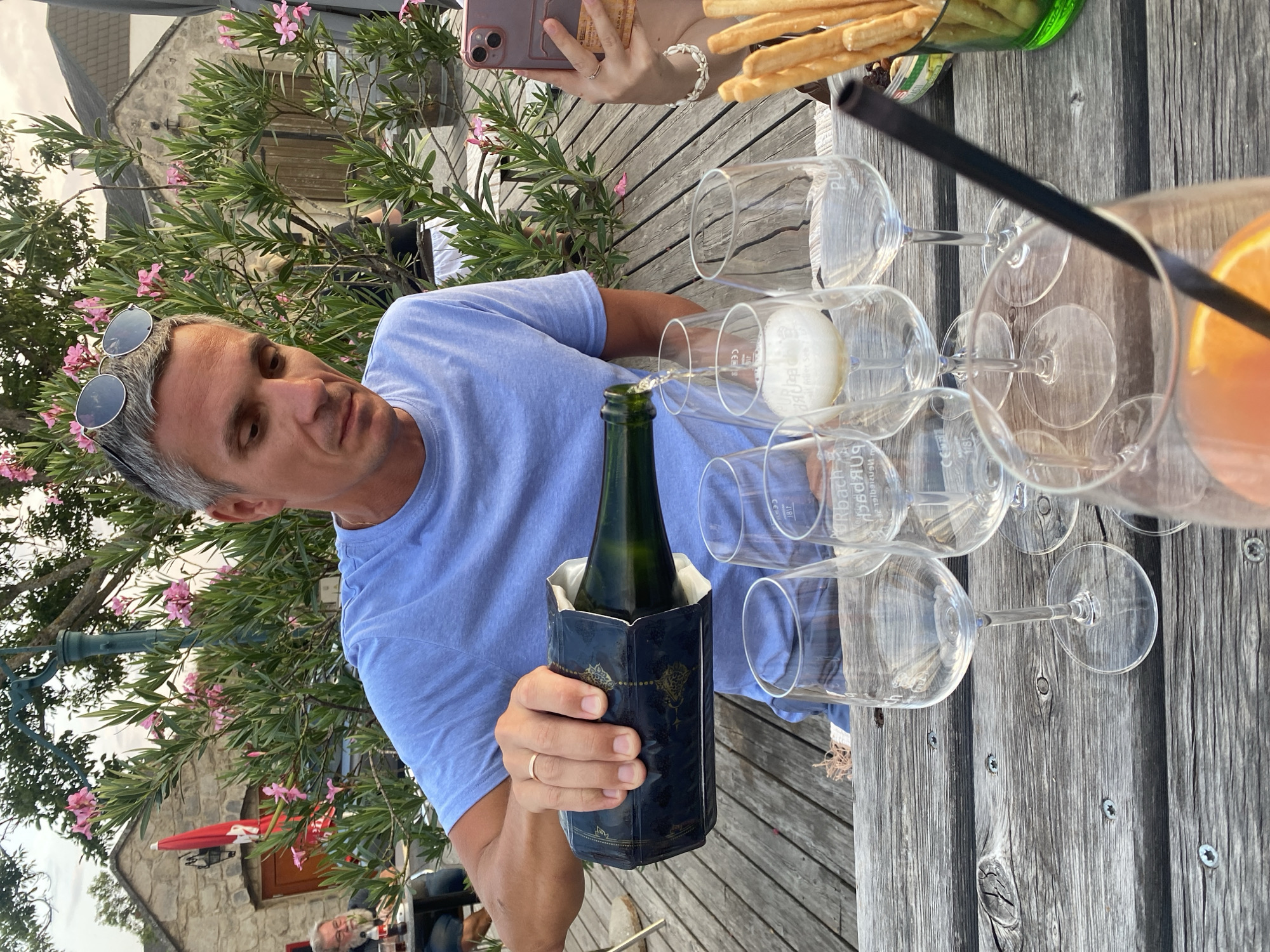In Purbach, Austria, a man with grey hair and sunglasses perched on his head is pouring a sparkling beverage from a dark bottle into wine glasses on a wooden table. The table is set with several empty wine glasses, one partially filled with the bubbly drink, and a glass containing an orange beverage with a black straw. A hand holding a pink smartphone is visible on the right, and a green container with breadsticks and a small dish with nuts is also on the table. In the background, a stone building with a red umbrella and pink flowers from a flowering bush are visible. Another person with a beard is seated at a table in the distance. The scene appears to be an outdoor dining setting.