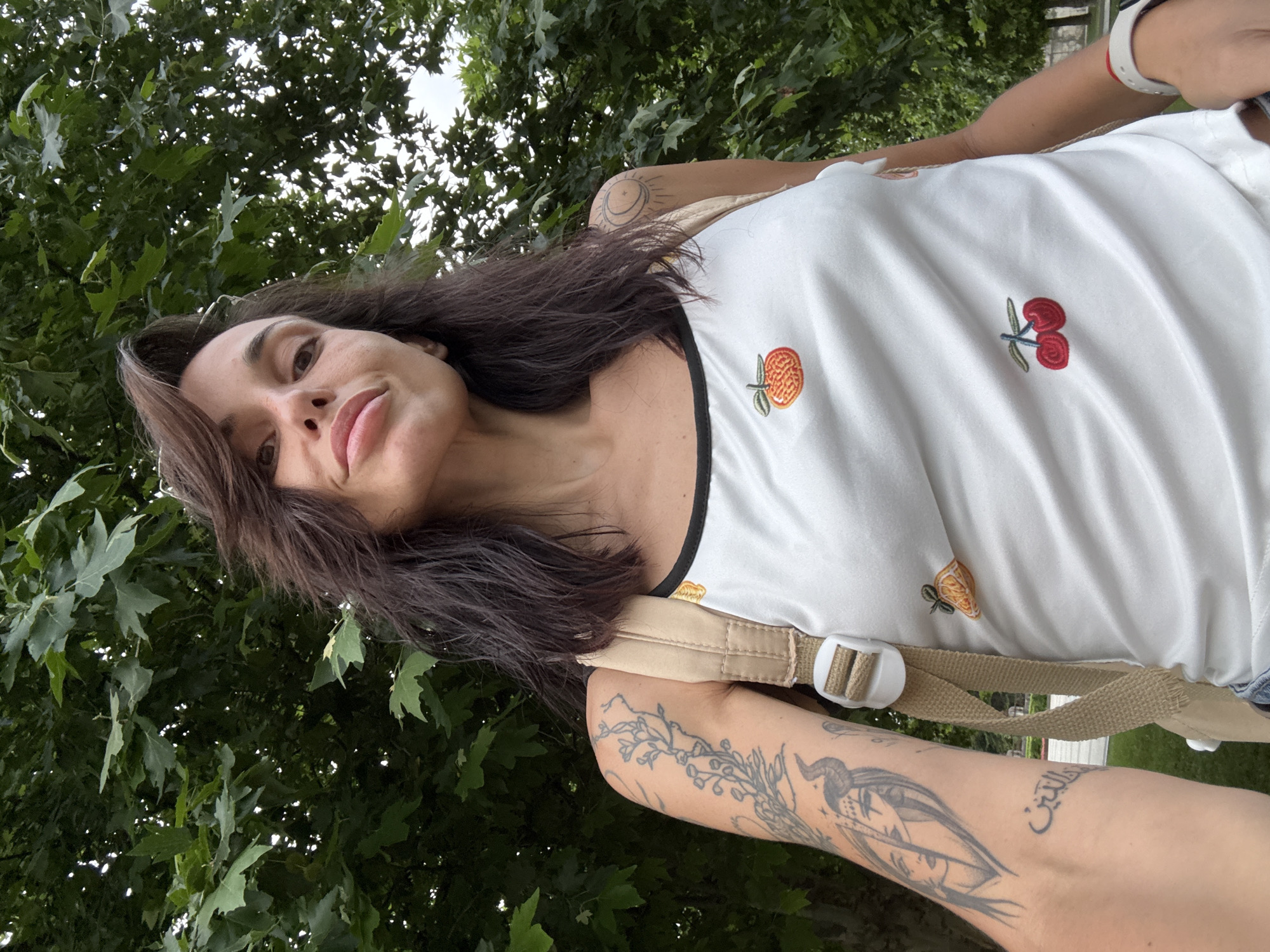An adult female is depicted in a close-up, upward-angled outdoor selfie. She has medium-length dark brown hair, fair skin, and full lips slightly parted. She is wearing a white tank top with a black trim neckline, adorned with embroidered fruit motifs: an orange centrally, an orange slice on her right upper chest, and two red cherries on her lower left torso. A light beige backpack strap is visible over her right shoulder, and a white watch is on her left wrist.
Visible tattoos include a floral design on her left upper arm, and on her left inner forearm, two female faces (one horned, one veiled) above Arabic script translating to "the religion" or "faith." A stylized crescent moon with a central dot is tattooed on her right shoulder.
The background features dense green foliage from large deciduous trees and patches of bright, overcast sky. Through a gap in the distant foliage, a paved path, low green hedges, and a classical stone column or monument are visible. The scene takes place within a park or green space in Pleven, Bulgaria.