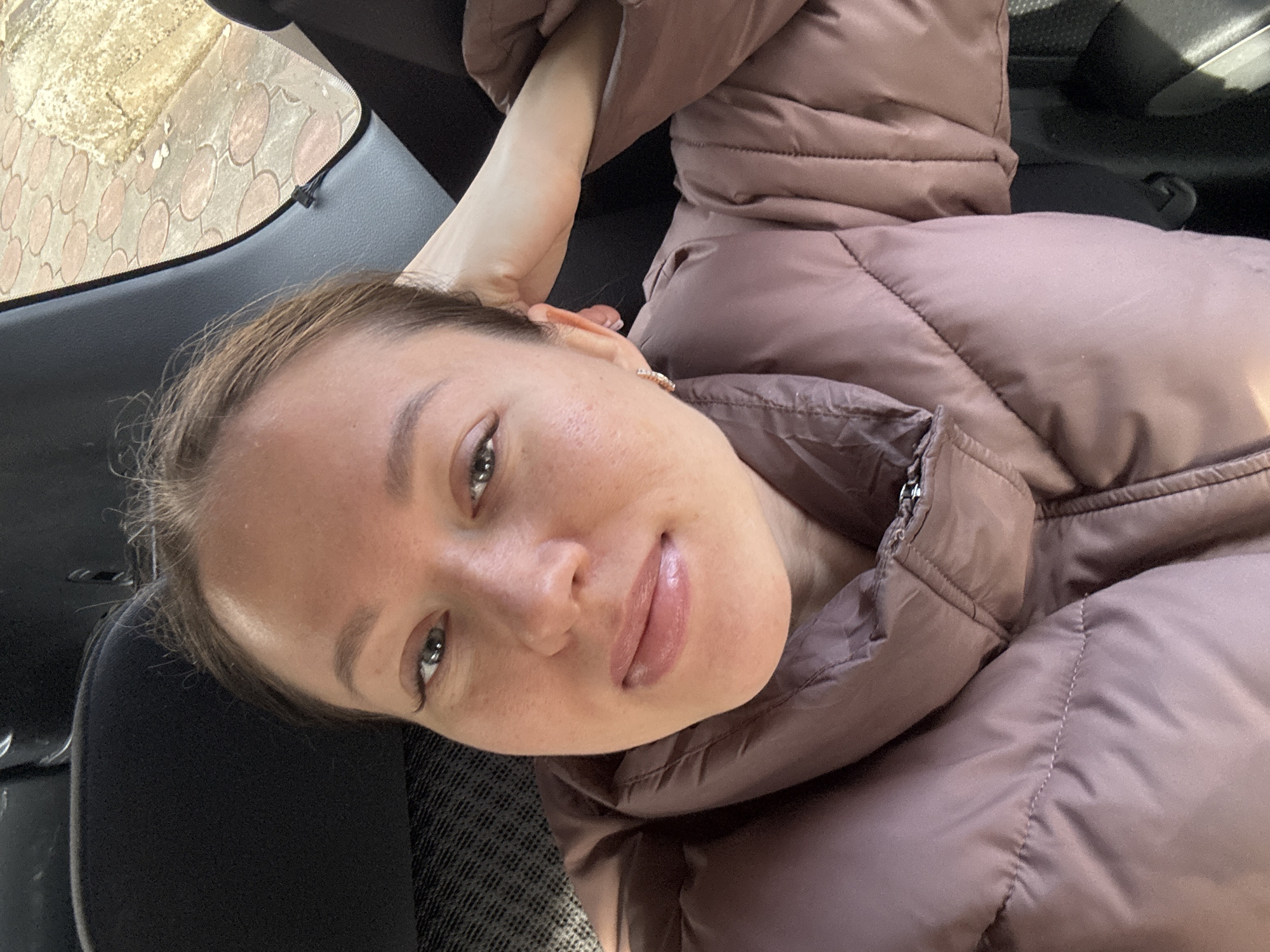 The image captures a selfie of a female individual seated within a vehicle in the city of Ферма, Russia. The woman, with light brown hair pulled back, fair skin, and natural makeup including mascara, looks directly at the camera with a slight smile. She wears a mauve-colored puffer jacket with visible stitching and a zipper collar. Her left arm is raised, with the hand resting behind her head, partially obscured by dark vehicle upholstery. A small, light-colored earring is visible in her right ear. The vehicle interior features dark grey fabric upholstery behind her head and a dark plastic trim piece along the right side of the frame. A black seatbelt strap is also visible over her right shoulder. Through the vehicle window, a section of outdoor ground is visible, paved with reddish-brown, possibly hexagonal or oval-shaped, interlocking stones, mixed with areas of lighter dirt or gravel. The scene is illuminated by natural daylight.