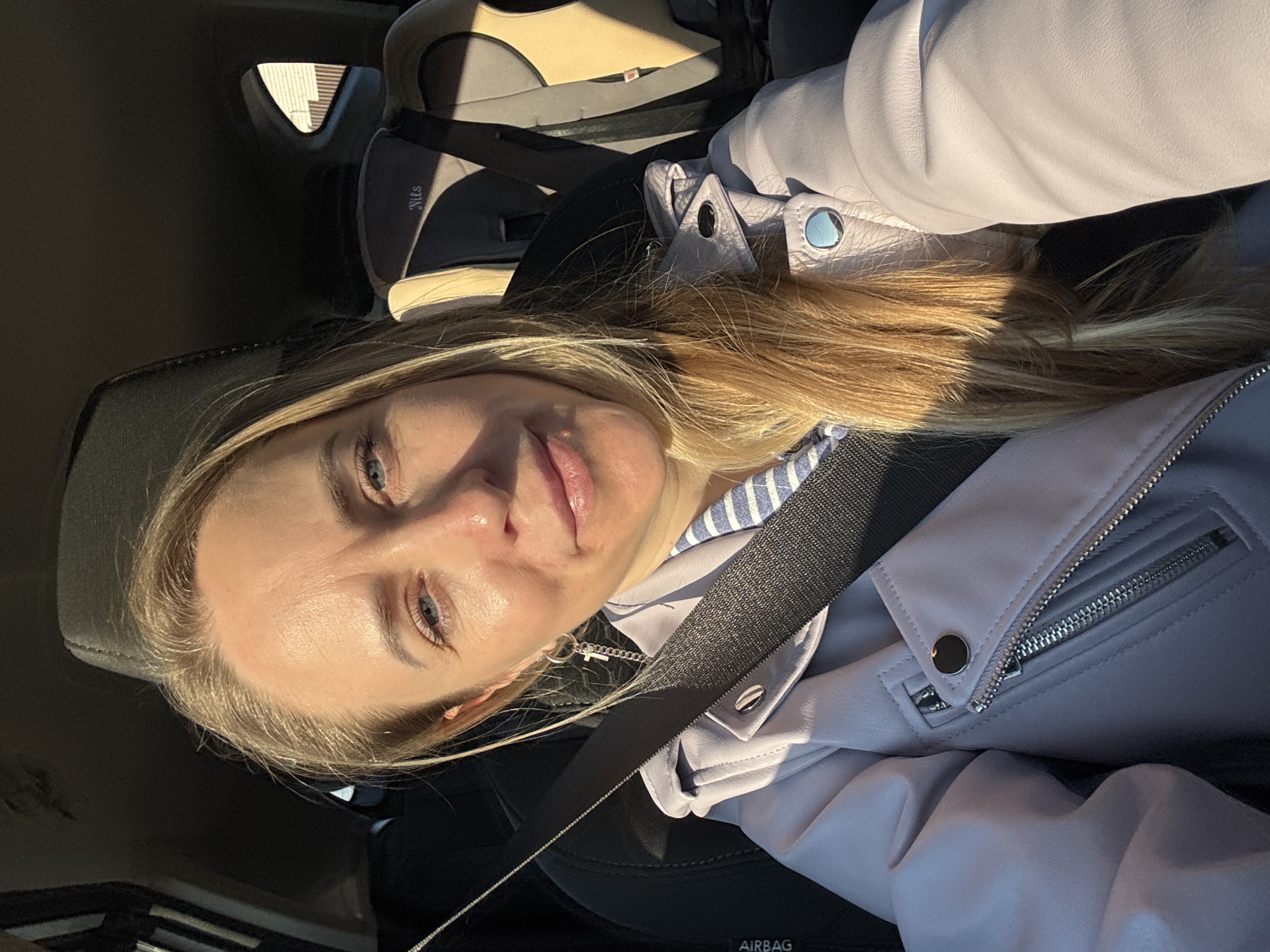 A blonde woman is sitting in the driver's seat of a car, wearing a lavender jacket and a seatbelt. A child's car seat is visible in the backseat. The location cannot be reliably determined to be Chaikovsky, Russia from the image.