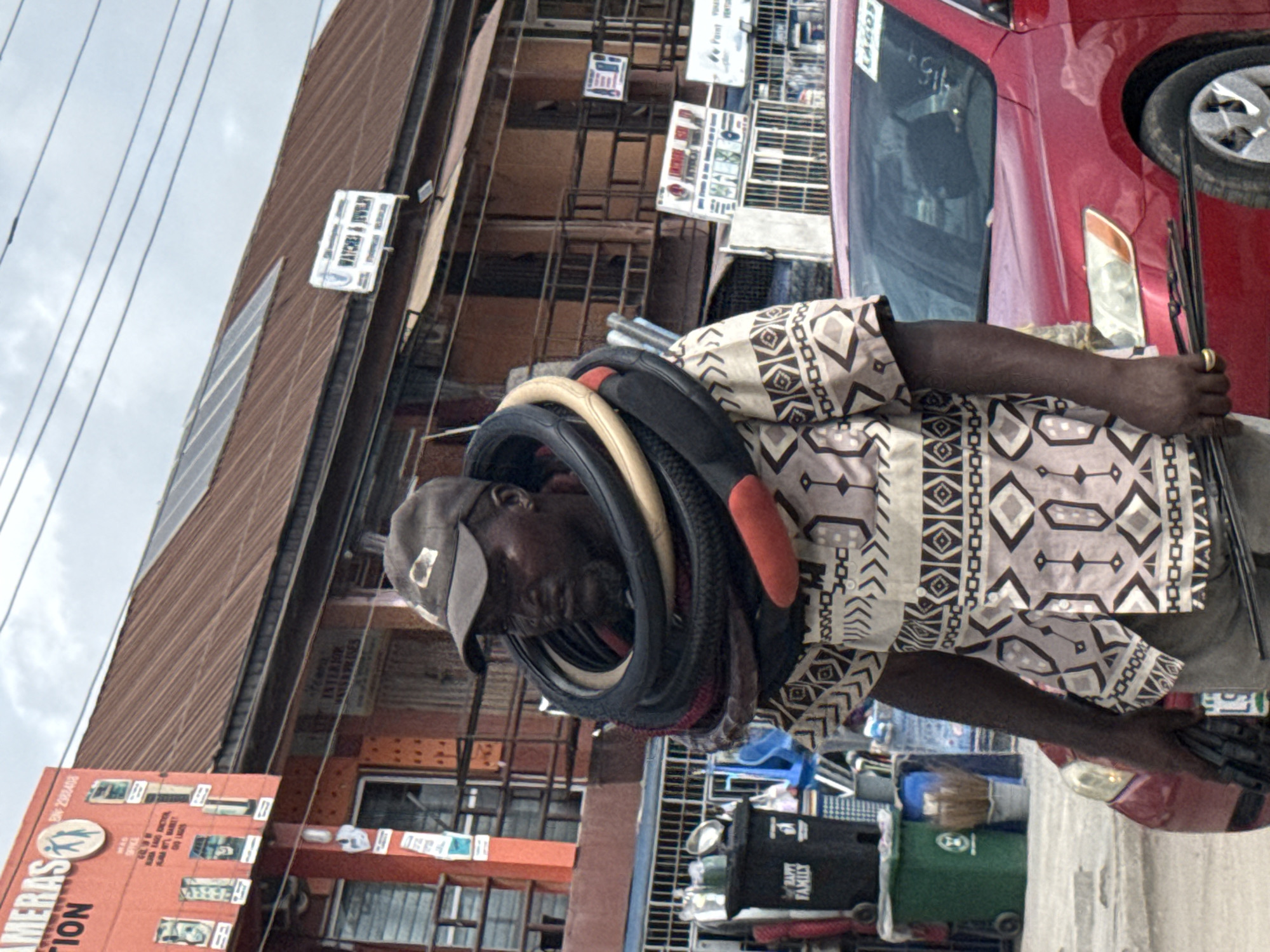 A man stands on a street in what appears to be Ado, Nigeria. The man is wearing a patterned short-sleeved shirt and a baseball cap. Around his neck is a stack of tire inner tubes. He holds multiple windshield wipers in his hand. The background includes multi-story buildings with signage, including "WATCHES & JEWELRY" and "LAGOS," and a car partially visible on the right edge of the frame. Waste bins are visible in the foreground.