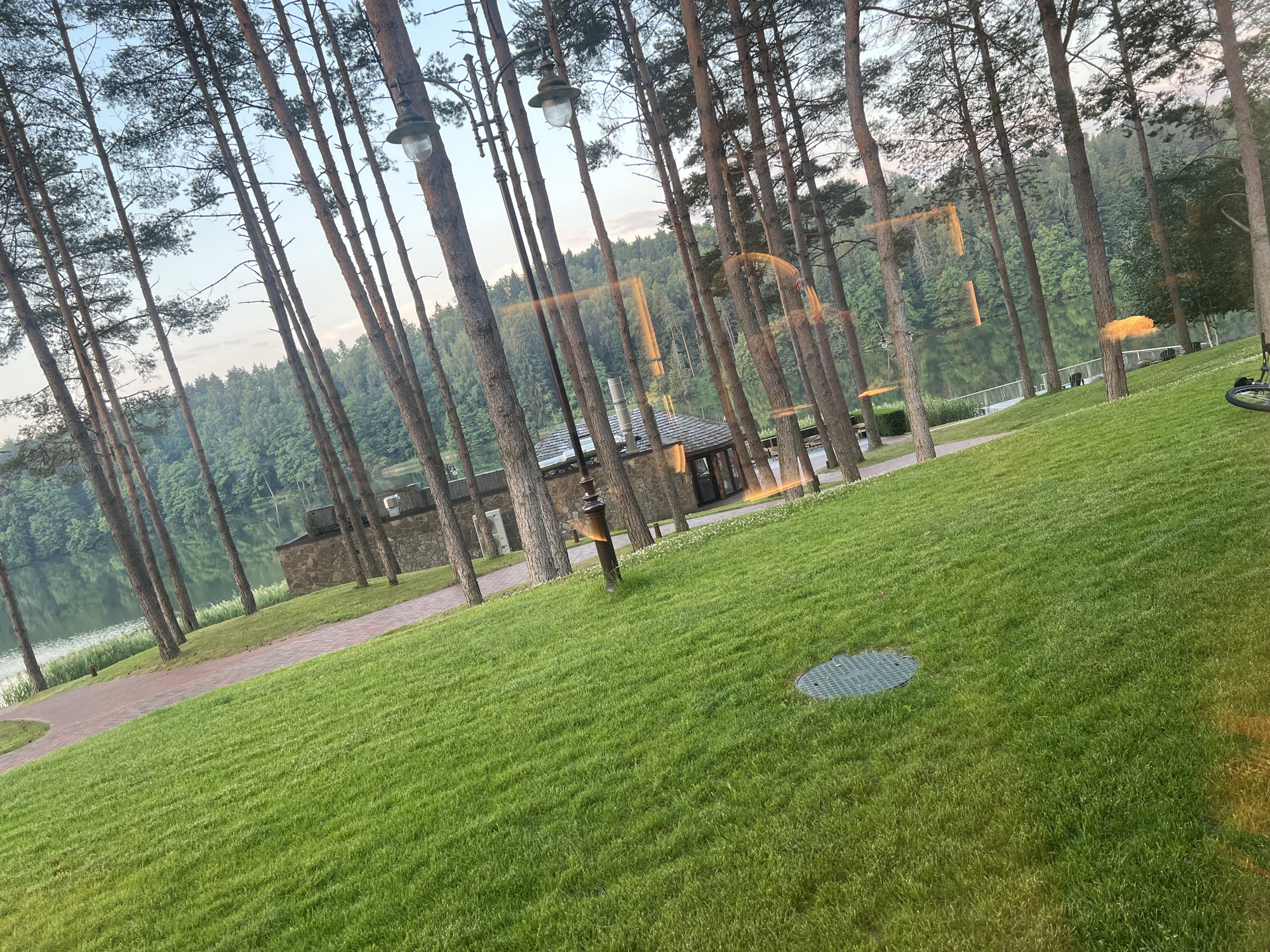 The image depicts a serene outdoor scene, likely a park or recreational area, situated near a body of water. Tall, slender pine trees dominate the foreground, their trunks creating a vertical pattern across the frame. A lush green lawn slopes gently upwards from the lower right to the mid-left of the image. A brick pathway curves through the lawn and leads towards a stone building with a dark-tiled roof. This building appears to be a small structure, possibly a pavilion or part of a larger complex, situated on the edge of a calm lake or pond visible in the background. The lake is surrounded by dense green forest, creating a natural backdrop. Several outdoor lamps are mounted on poles amongst the trees, suggesting the area is designed for evening use. The sky above is a pale blue with some scattered clouds. The overall impression is one of tranquility and natural beauty. The photographer's perspective is low, possibly from a seated position, and there are noticeable reflections on a glass surface in the foreground, indicating the photo might have been taken through a window. No people are clearly visible in the scene. The image does not provide explicit information about the city of Užiliai, Lithuania, other than it being the general location of the scene.