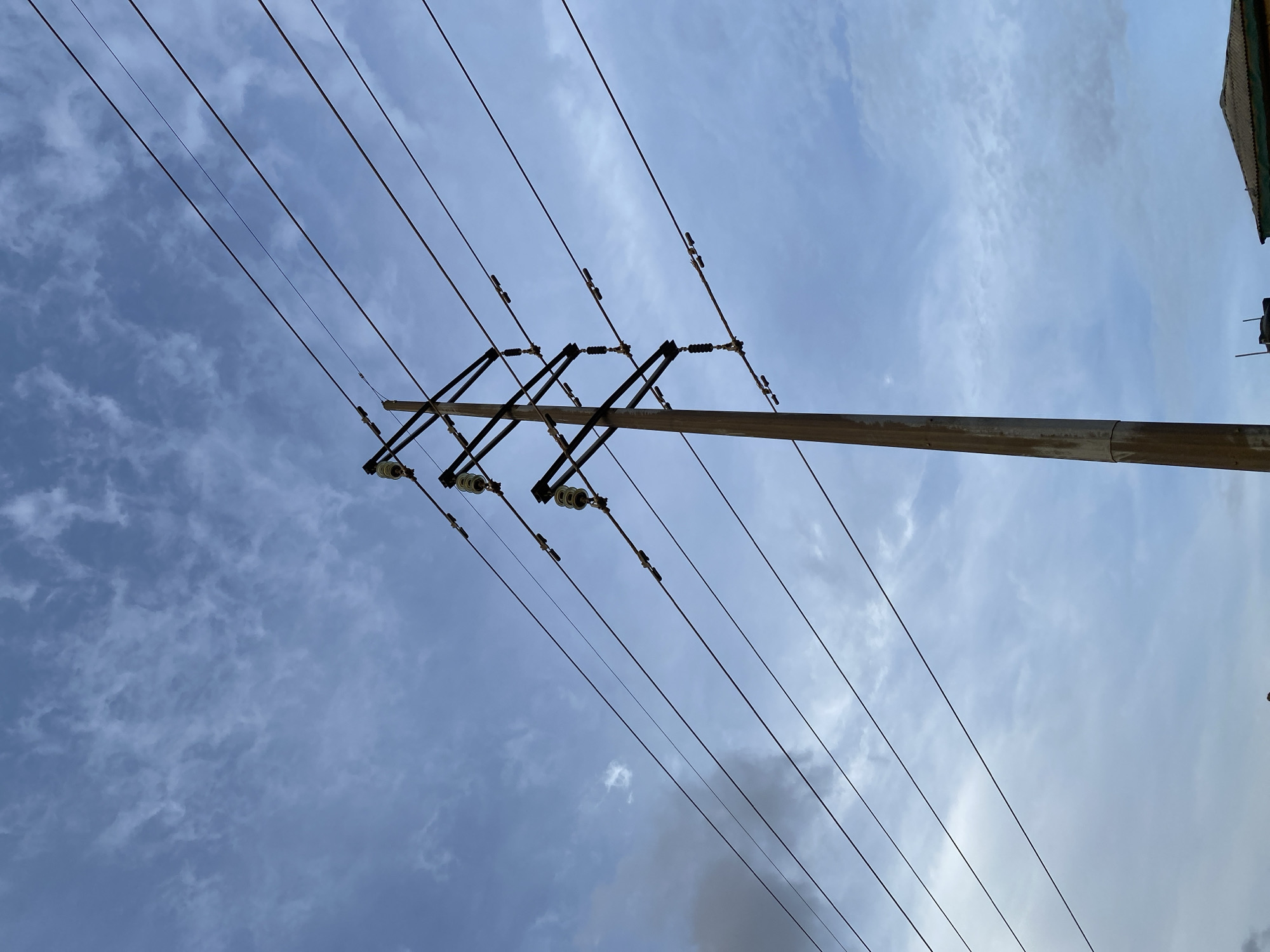 The image shows a low-angle view of utility poles and power lines against a cloudy sky. The utility pole is weathered, with black hardware suspending electrical insulators and wires. The sky exhibits varying shades of blue with textured cloud formations. The location is Lagos, Nigeria, based on visual characteristics of the utility infrastructure, the typical atmospheric conditions, and urban landscape features. There are no people visible. A section of the building at the top of the frame.