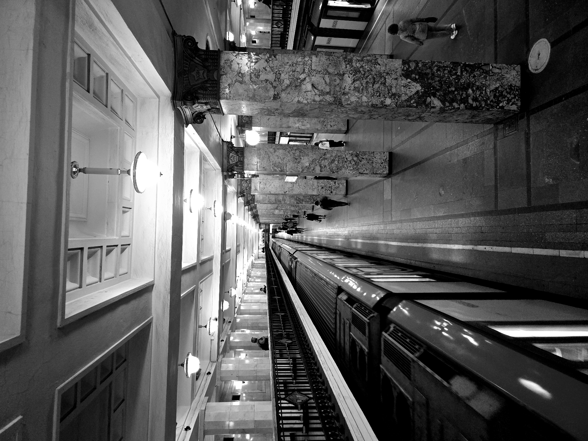 The image is a high-angle black-and-white photograph of the Mayakovskaya metro station in Moscow, Russia. The view is oriented lengthwise along the platform, with a train partially visible on the right side. The station's distinctive architectural features are prominent: white walls with gridded panels on the left, and rows of marble columns lining the platform in the center. People are walking on the platform. The image emphasizes the station's depth and grandeur.