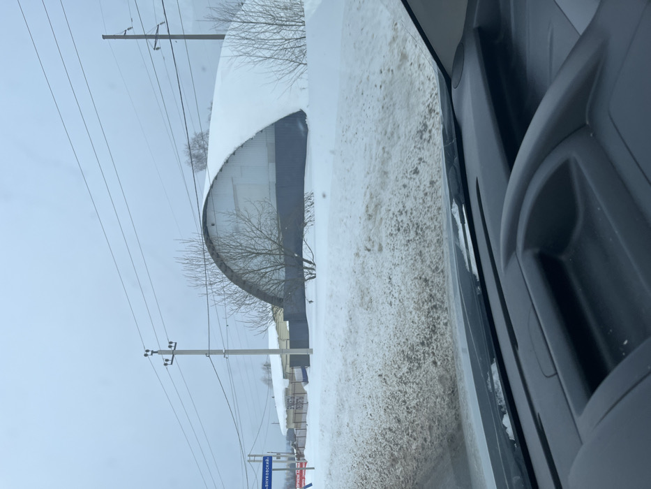The image captures a snow-covered winter scene in Poddubki, Russia, viewed from inside a vehicle. The lower frame reveals a dark gray vehicle dashboard and the base of the windshield, with minimal ice or snow visible along its edge. Directly outside, a substantial mound of dirty, brown-tinted snow occupies the foreground, suggesting roadside accumulation or slush.
Beyond this snow, a dark gray corrugated metal fence stretches across the mid-ground. Several bare deciduous trees and shrubs are scattered in front of and behind the fence, some partially covered by snow. The most prominent structure is a large, arch-roofed building, typical of a quonset hut, positioned to the right of the frame's center. This building features white or light gray corrugated metal paneling on its upper sections and a darker gray base, with its entire curved roof heavily blanketed in a smooth layer of white snow.
The background consists of an overcast, light gray sky. Multiple horizontal power lines are visible, supported by two concrete utility poles. To the far left, a blue road sign with white Cyrillic text, partially decipherable as "Г-ВНУКОВСКИЙ" (G-Vnukovsky), is visible above the snow line, with a red sign beneath it. Another white sign with black Cyrillic text, possibly indicating "БРИКЕТЫ" (Briquettes) and "КУПИТЬ" (Buy), is positioned behind the fence line, to the left of the arched building. No individuals, animals, or discernible actions are present in the scene. The environment is consistent with a wintry, semi-rural or industrial roadside setting.