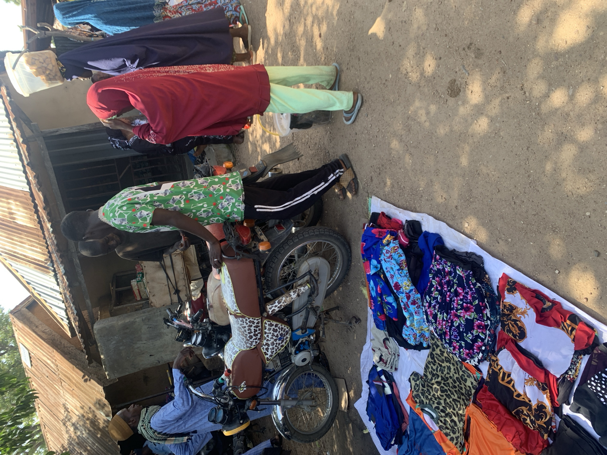 The image depicts an outdoor scene, likely a market, in Boi A, Nigeria. The ground is dirt, with sunlight casting dappled shadows, suggesting it is daytime. Several people are visible, mostly dressed in traditional African attire. One person, wearing a red garment and light green pants, appears to be attending to something on a motorcycle. Another individual in a blue shirt and yellow head covering is seated. The focal point includes several motorcycles and a spread of colorful clothing, including patterned dresses and scarves, laid out on a white cloth, implying a commercial activity or sale. The setting includes a corrugated metal structure on the left, possibly a shop or dwelling. The overall mood appears to be bustling and active, characteristic of a market environment. There is no discernible text in the image.