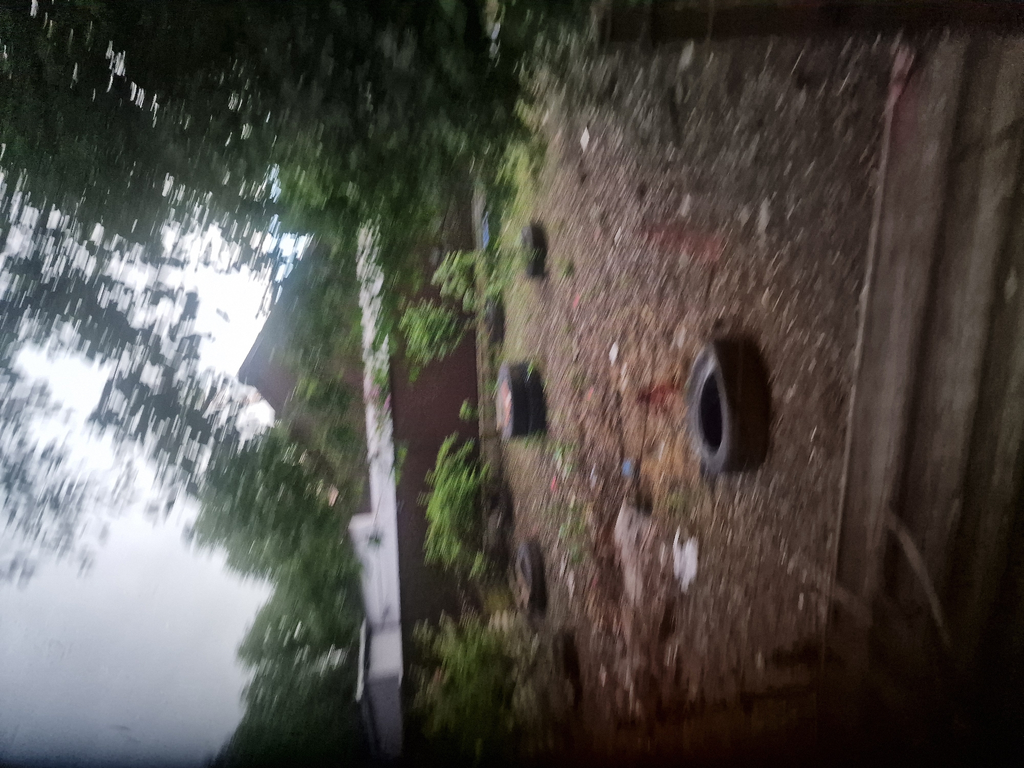 The image depicts an outdoor scene with several discarded tires scattered across a patch of ground covered with dirt and small debris. In the background, there is a building with a reddish-brown wall and a lighter colored roof, partially obscured by trees. The foliage is dense and appears to be a mix of deciduous trees. The sky is overcast, suggesting cloudy weather. The overall impression is one of an unkempt or abandoned area, possibly a backyard or a disused industrial space within the city of Kursavka, Russia, given the context of the prompt. The motion blur in the image suggests it was taken while moving, likely in a vehicle.