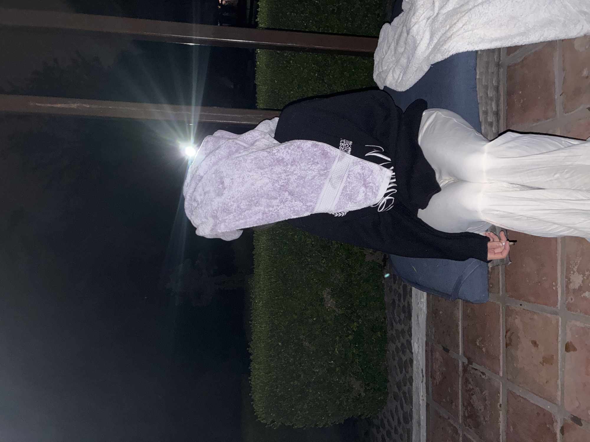A person is sitting on an outdoor sofa, their head covered with a purple towel. They are wearing a black hoodie and white pants. The sofa is topped with a white towel. The scene appears to be at night. The background includes a dark landscape with green bushes. The location is Cañada de Madero, Mexico.