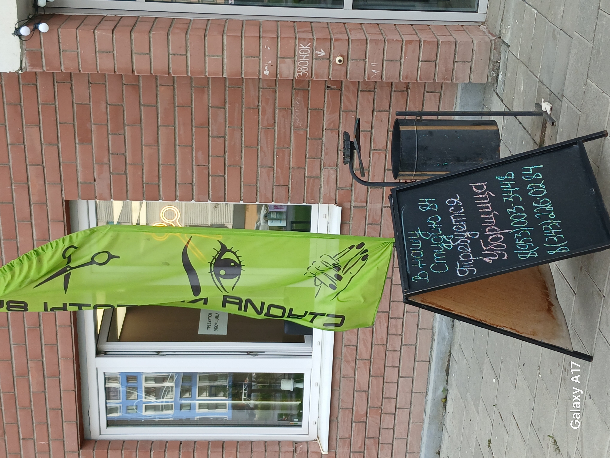 This outdoor, daylight shot depicts the facade of a brick building, likely a commercial establishment, on a paved sidewalk. The image is taken from a slightly low angle, showing a section of the red-brown brick wall with white mortar.
Prominently displayed is a bright green banner or flag, partially draped over a window. The banner features black stylized graphics: a pair of scissors on the left, an eye with long eyelashes in the center, and a hand with manicured nails on the right, suggesting a beauty salon offering hair, lash, and nail services. Through the window, parts of the banner and an internal sign are visible, reading "ТРЕБУЕТСЯ УБОРЩИЦА" (A Cleaner is required).
To the right of the banner, an A-frame chalkboard sign stands on the sidewalk. Written in chalk, it advertises a job opening: "В нашу Студию 84 Требуется Уборщица" (In our Studio 84, a Cleaner is required). Below this, two phone numbers are listed: "8(953)003-3448" and "8(343)226-0284". A black metal trash bin with a gold stripe is positioned behind the A-frame sign.
Above the window and to the right, a small handwritten sign on the brick wall points to a doorbell, stating "ЗВОНОК" (Ring/Call). A string of decorative white round lights runs along the top edge of the building facade. The pavement is made of gray interlocking pavers. The reflections in the window suggest other buildings across the street. The bright lighting indicates it is daytime. The scene appears to be on a city street in Yekaterinburg, Russia. The bottom right corner of the image contains a "Galaxy A17" watermark.