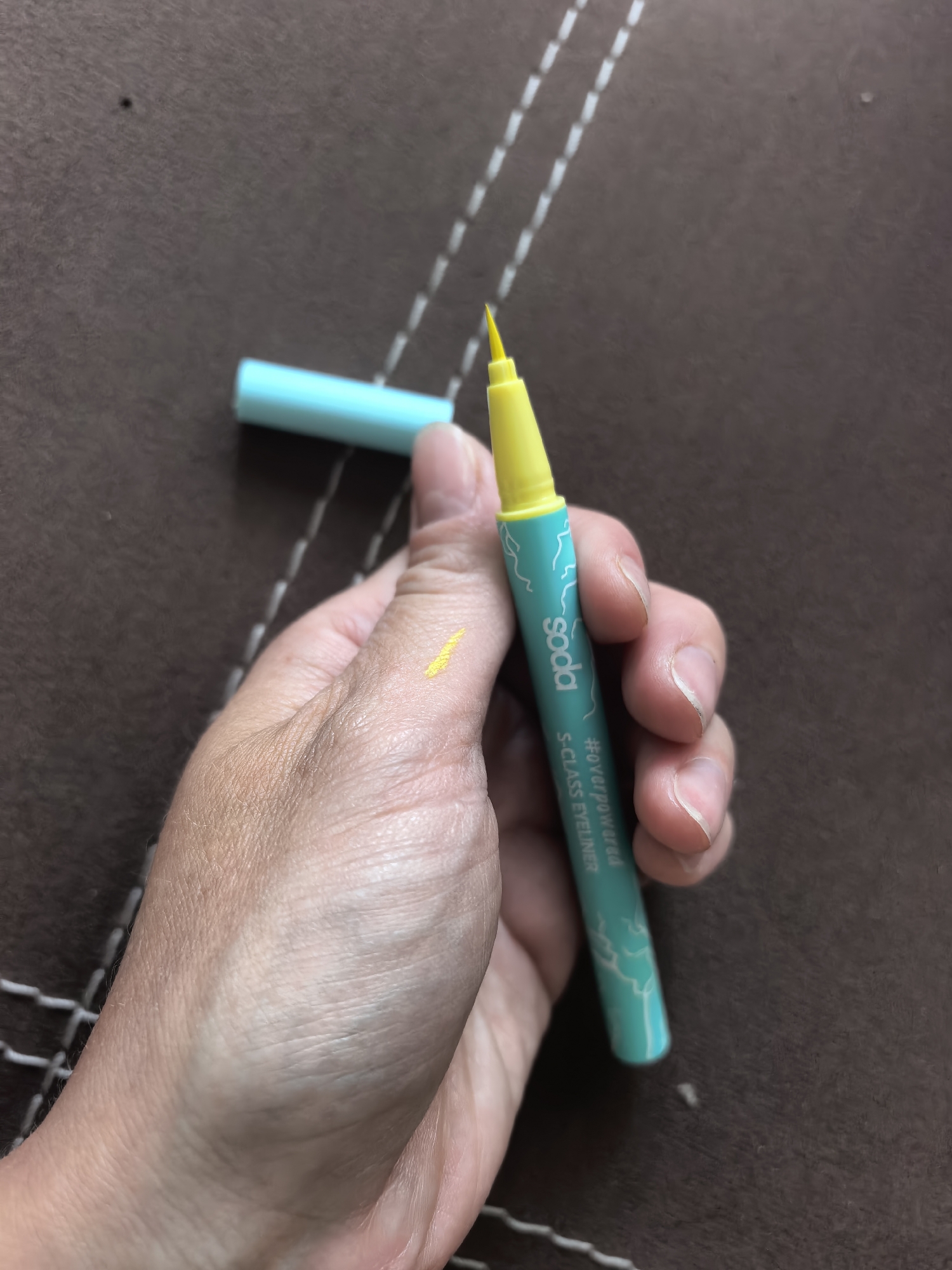 A hand holds an open yellow eyeliner pen, marked "soda #overpowereda S-CLASS EYELINER" with a turquoise body, on a brown textured surface. There is a yellow streak on the back of the hand. The pen cap is located nearby. The location in the city of Sterlitamak, Russia is not discernible.