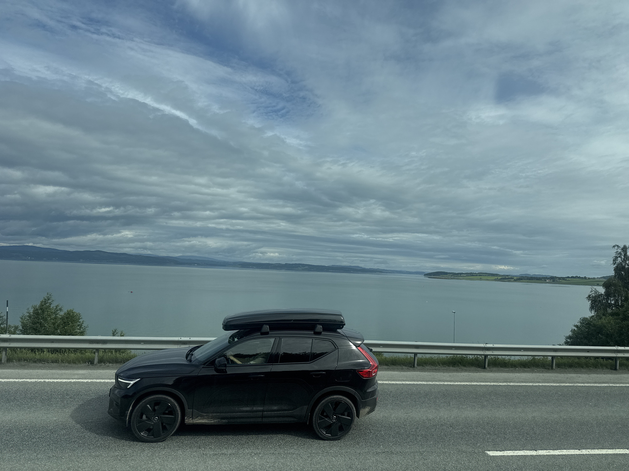 A black Volvo XC40 SUV with a black cargo box on its roof is driving on a paved road in Skogn, Norway. The car is black and has black wheels. The road has white lane markings. A metal guardrail is visible to the left of the road, with grass and trees behind it. Beyond the guardrail, a large body of water, likely a fjord, stretches to the horizon. Hills and distant land are visible across the water. The sky is cloudy and overcast. The image appears to have been taken from another vehicle moving in the same direction.
