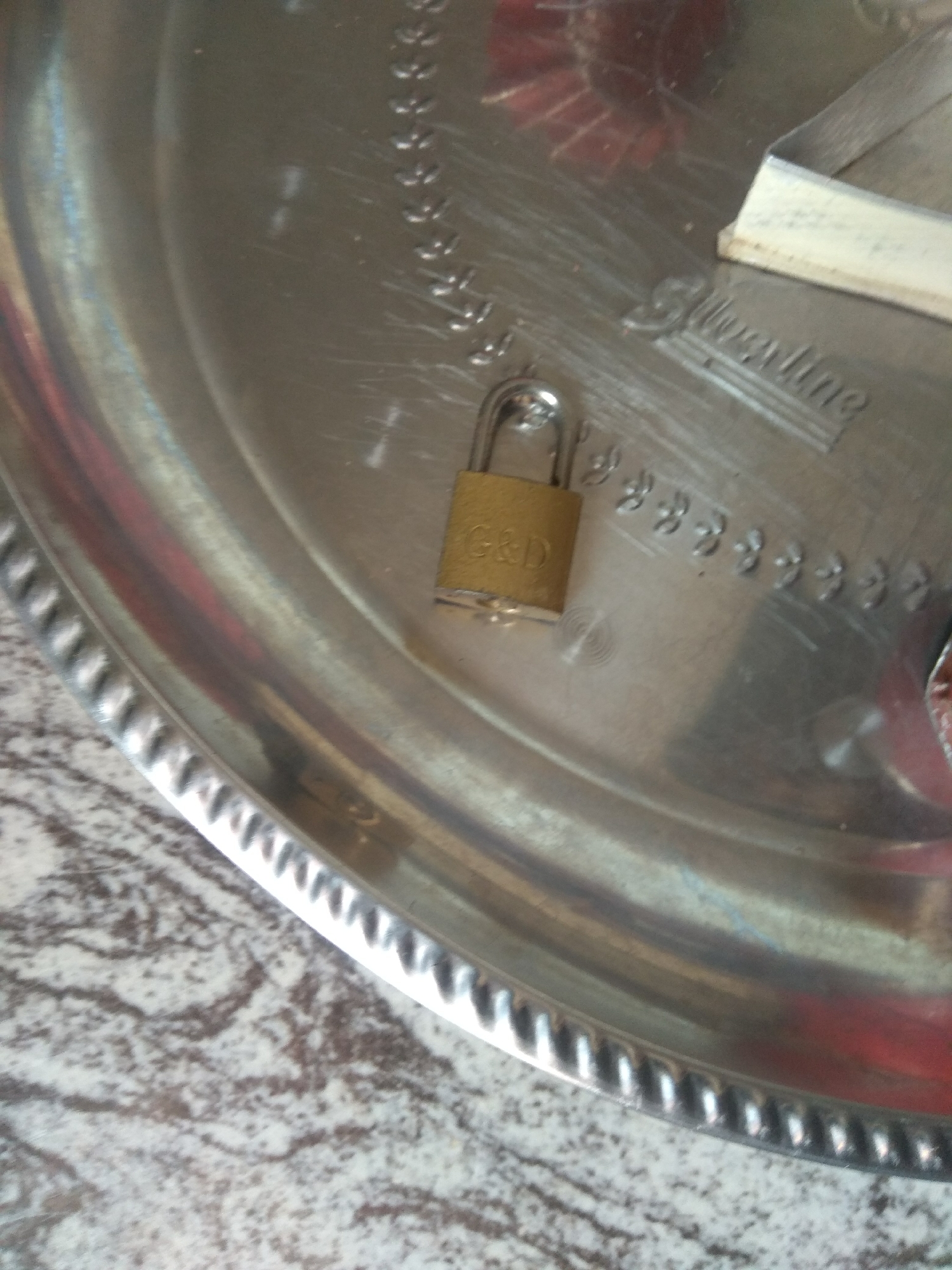 A close-up, overhead shot of a small, brass-colored padlock is centered on a silver-toned tray with a decorative rim. The padlock is oriented vertically with its shackle facing upwards and is inscribed with the letters "G&D" in black. The tray is resting on a speckled surface that appears to be a marble countertop or table. The tray has a subtle engraved pattern of repeating floral or decorative motifs. In the background, a white object with a textured surface is partially visible, and a red-colored object with a radial pattern is also present. The lighting suggests an indoor setting. There are no people visible in the image. The location is not identifiable as Jalingo, Nigeria.