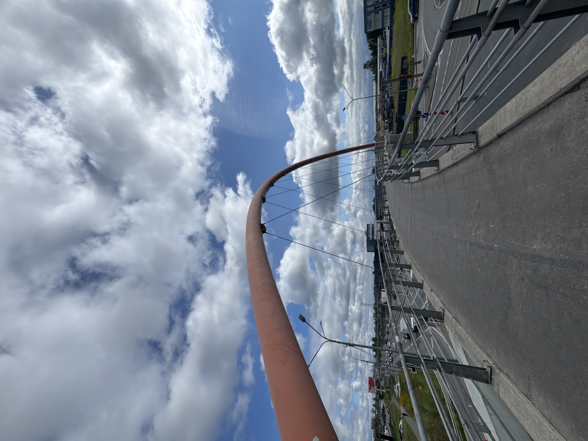 The image shows a wide, pedestrian overpass with a distinctive arched red structure overhead. The overpass has metal railings on both sides. Below the overpass, a multi-lane road with moderate traffic is visible, featuring cars and trucks. The sky is partially cloudy with scattered white clouds against a blue background. In the distance, buildings and trees are visible, suggesting an urban or suburban environment. There are no people clearly visible on the overpass in the main shot. The overpass appears to be part of a modern infrastructure project, possibly connecting different parts of the city or providing safe passage over a busy road.