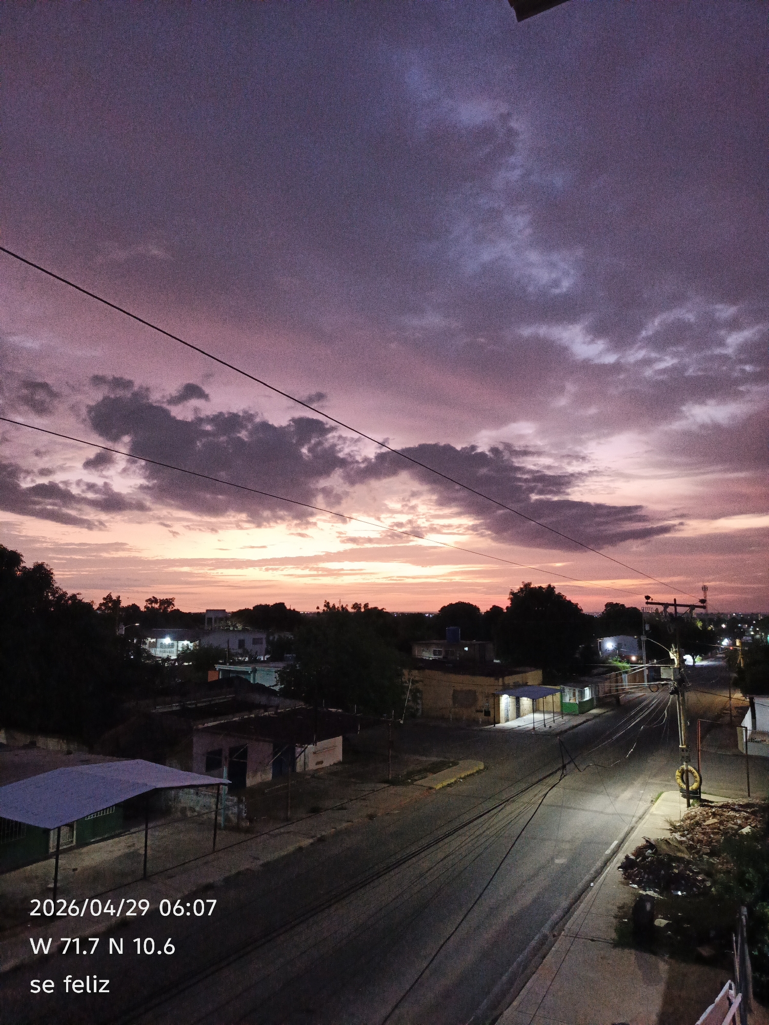 Here is a detailed description of the image:
**Environment:**
The image shows an urban environment, seemingly a residential area, under a dramatic twilight or sunset sky. The time and location are recorded at the bottom left: "2026/04/29 06:07, W 71.7 N 10.6," which points to a location near San Francisco, Venezuela.
**Sky:**
The sky is the most prominent feature, with a blend of purple, pink, and orange hues. There are scattered clouds that create a dynamic and textured appearance.
**Landscape:**
The cityscape is visible in the middle ground. Buildings are mostly single-story or low-rise structures. Streetlights illuminate the area, creating a network of lit pathways.
**Objects:**
- Overhead power lines run across the sky.
- A street with paved roads and sidewalks.
- A utility pole is on the right side with a ring around it.
**Text:**
The phrase "se feliz" ("be happy" in Spanish) is written at the bottom left corner.