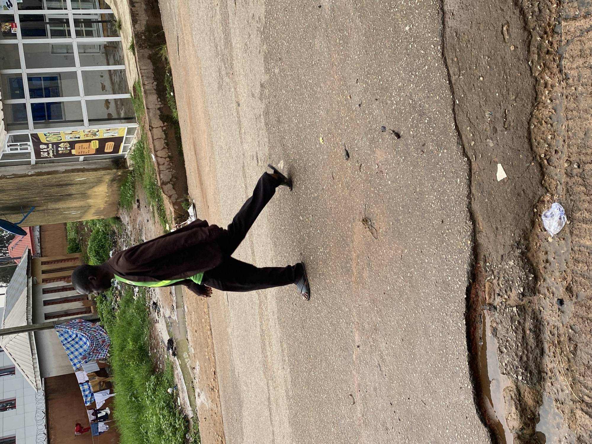 In Jos, Nigeria, a Black man wearing a brown blazer over a bright green shirt and black pants walks on an asphalt road. He is wearing open-toed sandals. To his left, clothes hang on a clothesline in front of a building. The road shows signs of disrepair, with a large pothole filled with water and debris. In the background, another building with large windows and a sign that reads "EAT & SIP" is visible. The scene appears to be an urban street with evidence of daily life and infrastructure challenges.