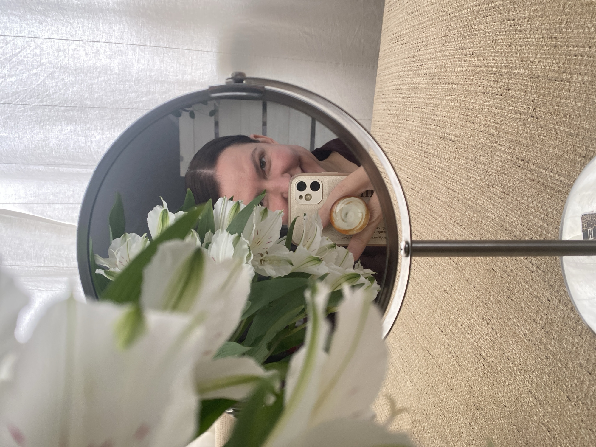 A selfie taken in a round mirror shows a woman holding a gold iPhone with a pop-up grip on the back. She is positioned to the left of the mirror, with her face partially visible as she captures the reflection. A bouquet of white and green Alstroemeria flowers is in the foreground, partially obscuring the mirror's reflection. The mirror is mounted on a wall with beige textured wallpaper. A white, slatted window covering is seen in the background. There is no verifiable evidence in the image to support the claim that this scene is in Chaikovsky, Russia.