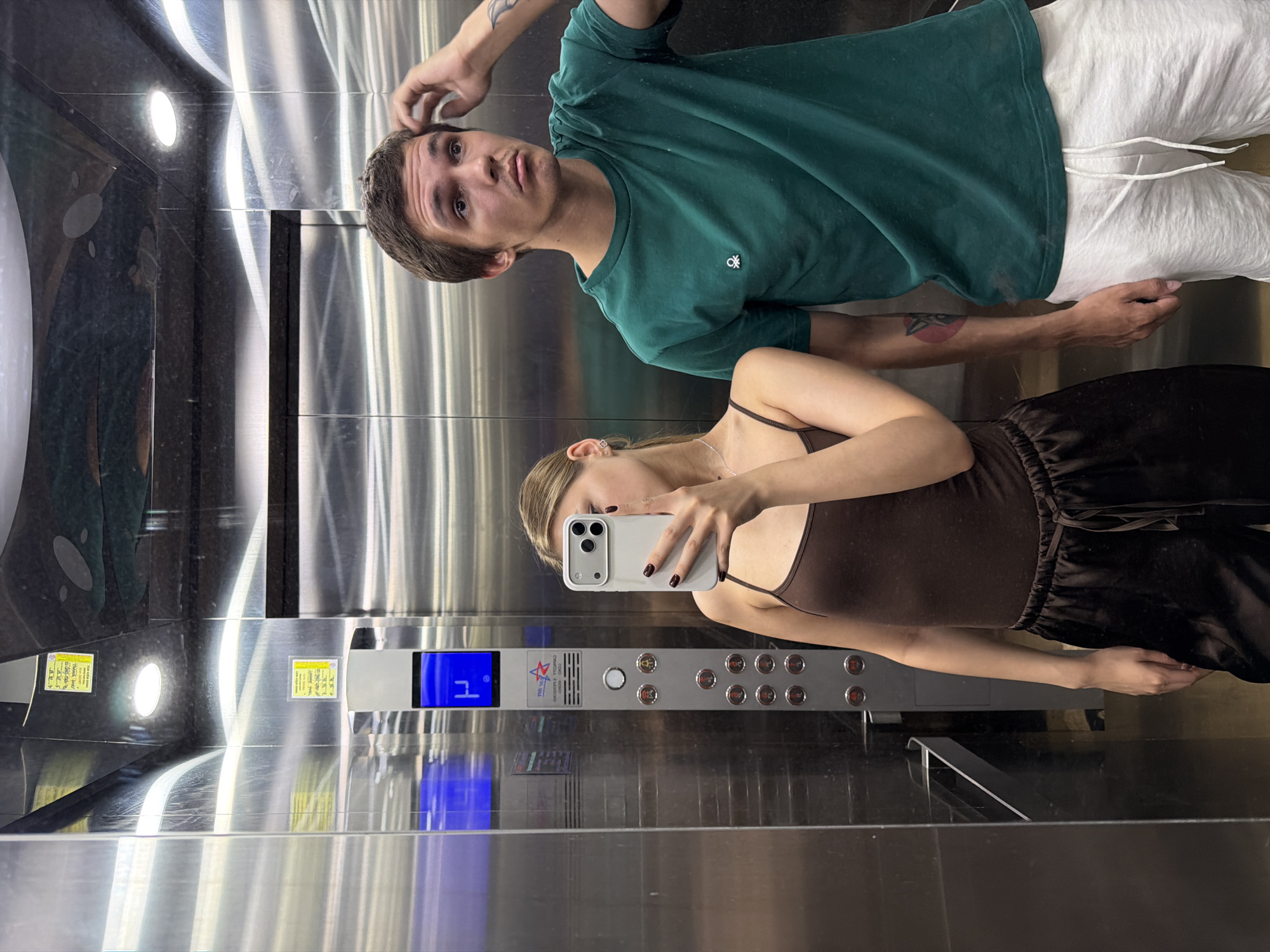In an elevator, a young man and woman are taking a mirrored selfie. The man, wearing a green t-shirt and white shorts, has his hand on his head, looking up and to the right. The woman, in a brown tank top and dark pants, holds a phone in her right hand, capturing the reflection. The elevator's metallic walls reflect the interior, including the illuminated floor indicator displaying "P" and a row of buttons. Two yellow stickers with Vietnamese text are visible on the wall above the control panel. The scene is set within a modern building, likely in Đà Nẵng, Vietnam, as indicated by the presence of Vietnamese text on the stickers.