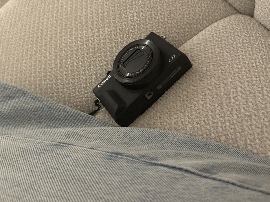 A black Canon G7X camera rests on a textured, cream-colored surface, likely a sofa or cushion. The camera's lens is prominent, displaying technical specifications. A portion of a light blue denim garment, possibly jeans, is visible in the lower left corner of the frame, creating a casual, relaxed setting. The composition is close-up, emphasizing the camera and its surroundings.