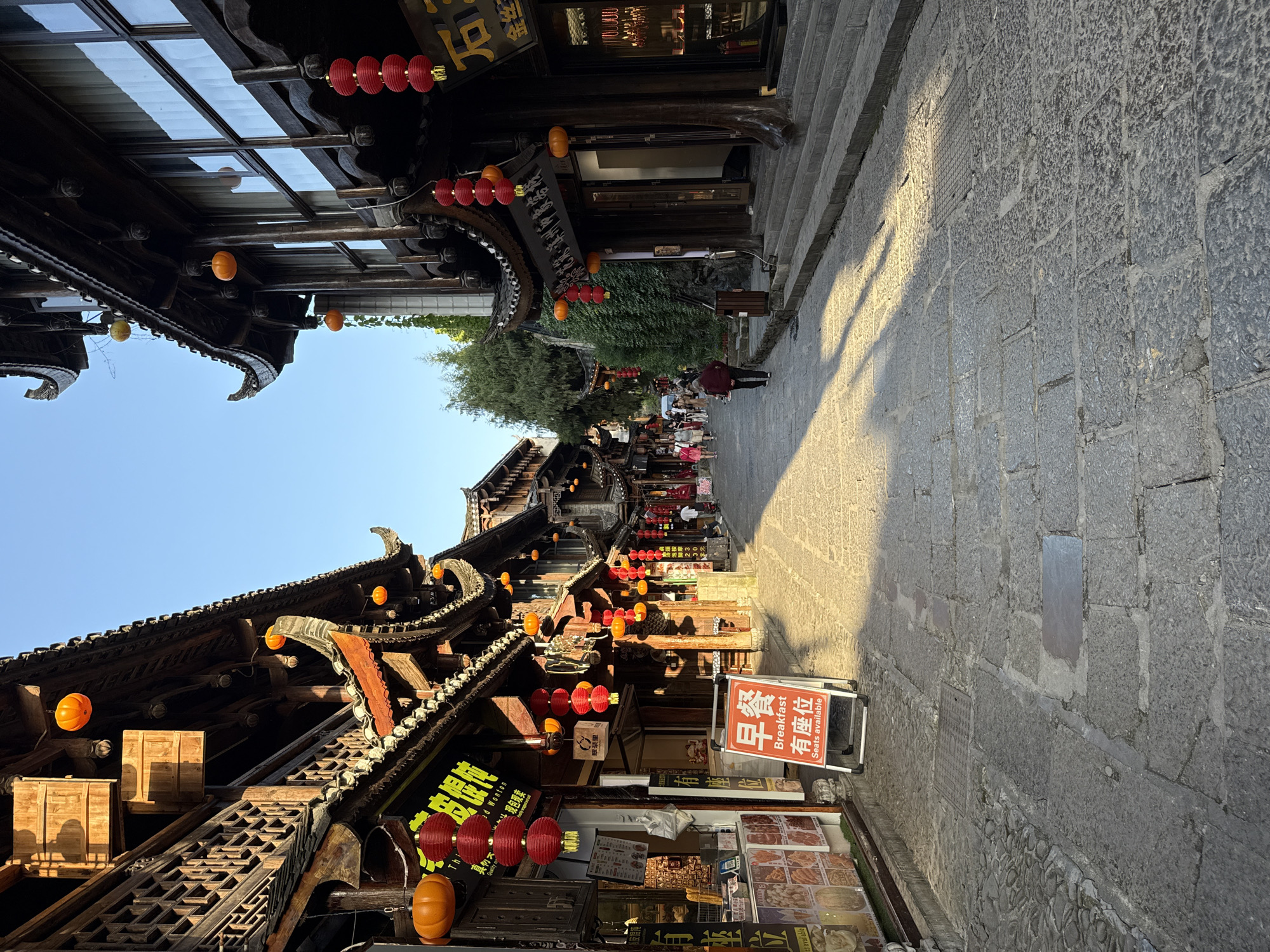 The image depicts a street scene in Furong, China. The architecture features traditional wooden buildings with ornate roofs, decorated with red and orange lanterns. A sign indicates "Breakfast, Seats Available" in both Chinese and English. People are seen walking in the distance. The street is paved with stone. Shopfronts display various items for sale.