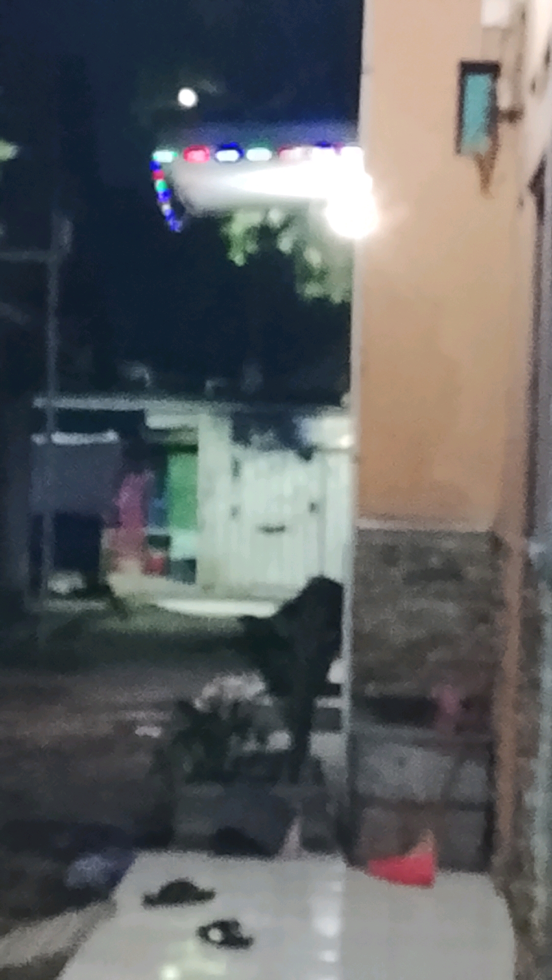 The image depicts a nighttime scene, likely in an outdoor area of Ujungrusi, Indonesia, possibly a yard or alley. In the foreground, a white surface, perhaps a table or bench, is visible with two dark objects on it, resembling footwear. To the right, a portion of a building with a light-colored facade and a darker, textured base is present. Further into the background, a dark, shadowy figure, possibly an animal or a person, is partially obscured by a wall. The background shows a dark environment with some illuminated structures and what appear to be colorful lights in the distance, suggesting a festive or populated area. A single bright light source is also prominent in the upper left quadrant of the image.