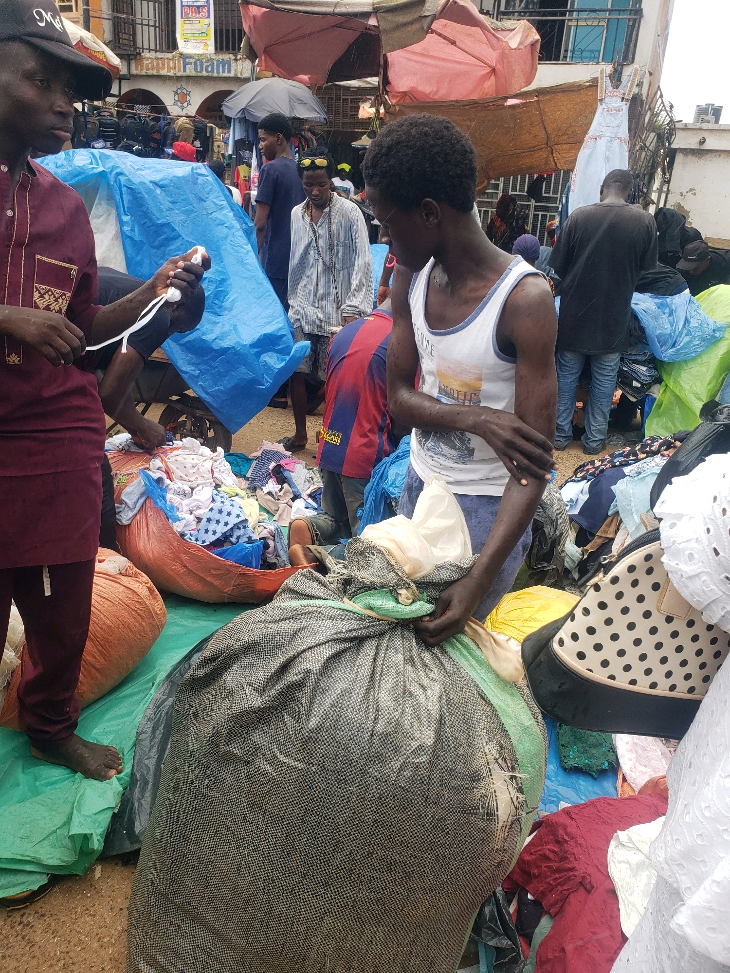 In Sabon Tasha, Nigeria, a busy marketplace is depicted with a focus on the foreground where two young men are engaged with large piles of clothing. One man, wearing a maroon dashiki and a black cap, is holding a white object in his hands. The other man, in a white tank top, stands beside him, with his arms visible. They are surrounded by various clothing items, some neatly folded and others in piles, covered by blue and green tarpaulins. In the background, a diverse group of people are seen amidst stalls displaying merchandise. The overall scene suggests a vibrant and bustling commercial environment, likely during a daytime market operation.