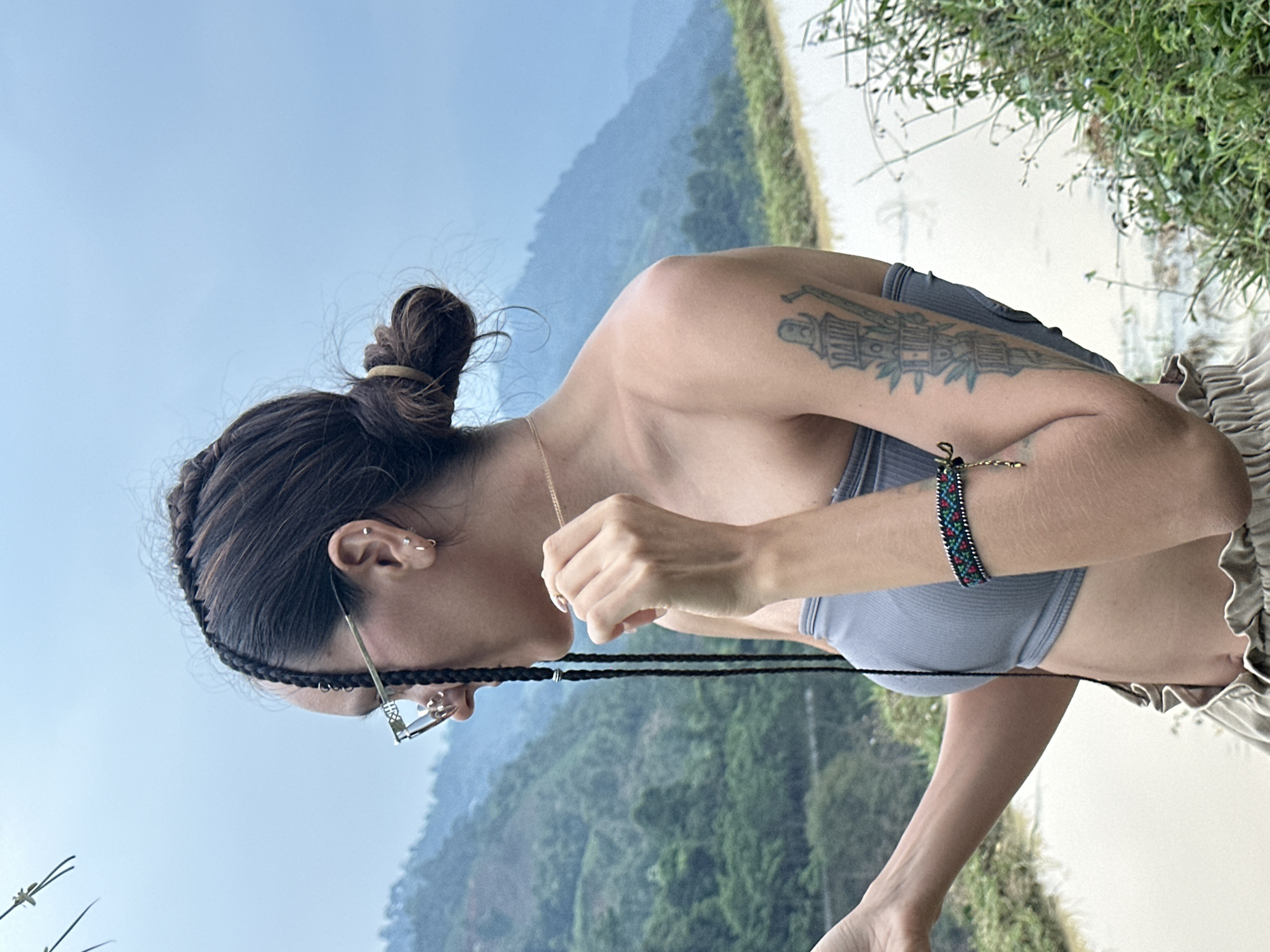 The image captures a side profile of a woman standing outdoors in Nậm Dịch Commune, Vietnam. She is looking downwards and slightly to her left. Her dark hair is styled with intricate braids framing her face and gathered into a high bun on top of her head, secured with a light-colored hair tie. She wears clear-rimmed glasses and multiple silver ear piercings, including hoops and studs. A thin gold chain necklace is visible around her neck.
She is wearing a grey, ribbed crop top, exposing her midriff, and light-colored, possibly beige, ruffled shorts or a skirt is visible on her right side. On her left upper arm, she has a detailed tattoo depicting a structural or architectural design. Her left wrist is adorned with a colorful, beaded bracelet featuring red, green, and black patterns, alongside a delicate gold bracelet. Her right hand is positioned near her chest/neck area, with fingers slightly curled.
The background features a serene natural landscape. Behind her, lush green mountains rise under a clear, light blue sky, indicating daytime and good weather. Below the mountains, a lighter area suggests a body of water or distant terrain. To her immediate right, vibrant green foliage and sparse branches are visible, framing the scene. No visible text is present in the image.