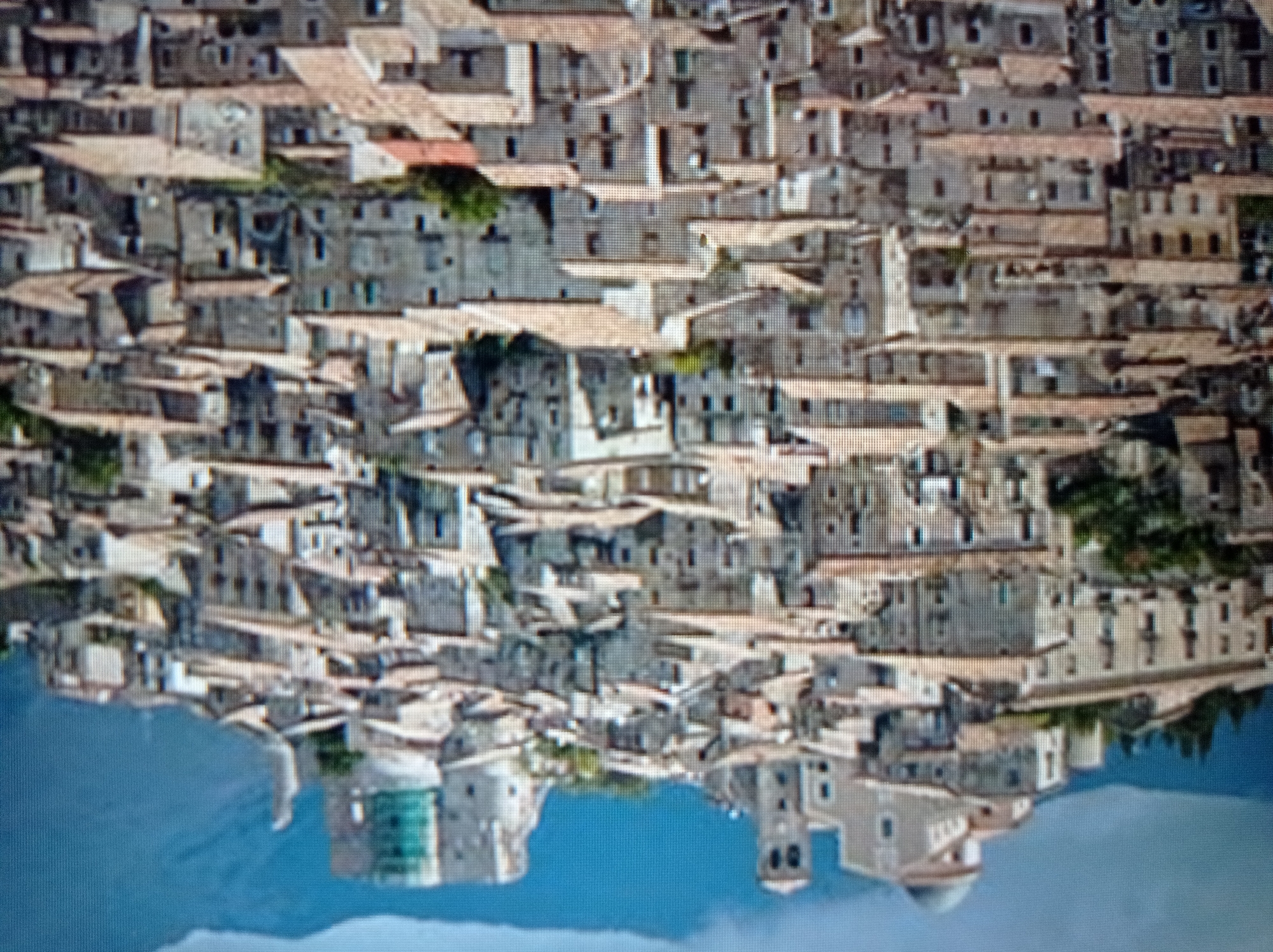 The image is a panoramic view of a dense, historic town perched on a hillside, with mountains in the background. The architecture consists of numerous stone buildings with terracotta tiled roofs, tightly packed together, creating a layered effect as they ascend the slope. Several prominent structures stand out: a large building with a dome and a bell tower on the left, and a fortified structure with a cylindrical tower on the right. The sky is a gradient of blue, with some darker, cloud-like formations at the top. Vegetation, including trees and bushes, is visible interspersed among the buildings, particularly on the slopes. The overall impression is of an ancient European settlement. The image does not show the city of Mukachevo, Ukraine, or any recognizable landmarks from that location.
