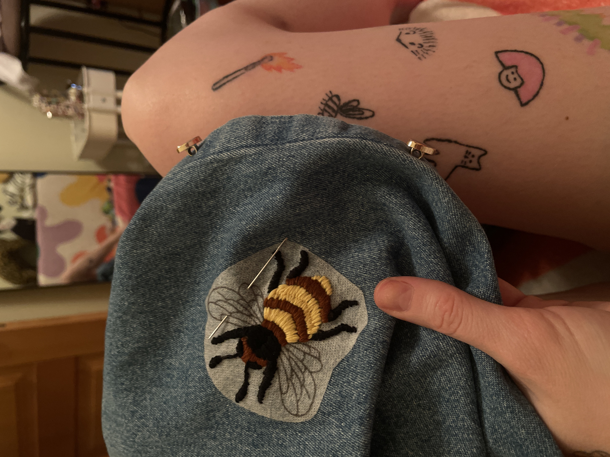 The image captures a close-up, high-angle view of a person engaged in a crafting activity, likely embroidery or appliqué, in an indoor setting in Belgrade, Serbia. The primary subject is a denim garment, possibly a sleeve or pant leg, to which an embroidered bee patch is being attached.
The bee patch features a detailed bumblebee design with a striped body in yellow and brown threads, a dark brown head, and black legs. Its wings are outlined in gray thread on a light gray fabric base. Two silver pins are visible, securing the patch to the denim. A light-skinned hand, with a thumb visible on the right, holds the denim garment.
Above the denim, a section of the person's light-skinned arm or leg is visible, adorned with several small, minimalist tattoos. These include a burning matchstick with an orange flame, a black outline of a winged insect (possibly a moth or bee), a small figure with spiky hair (resembling a hedgehog or a stylized person), a smiling face with pink, rounded hair, and a black outline of a cat-like figure.
The background is out of focus, showing blurry shapes and colors that suggest domestic furniture with wooden edges and possibly patterned fabrics or decor, indicating an indoor environment. The lighting appears to be artificial, consistent with an indoor evening or daytime setting with diffused light. There is no visible text in the image.