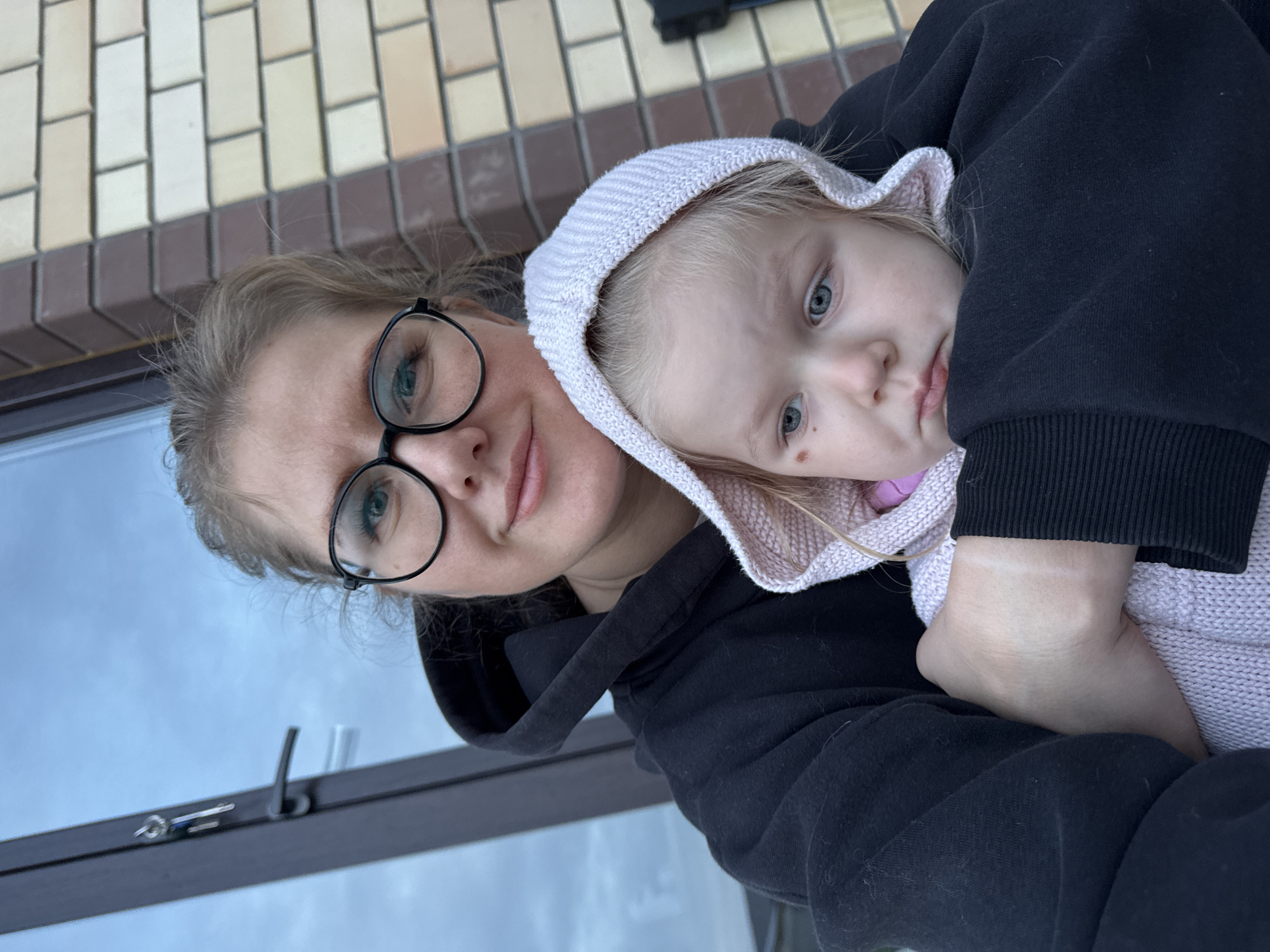 A woman wearing black glasses and a black hooded sweatshirt is holding a young child wearing a light pink hooded sweater. The woman appears to be smiling slightly. The child has blonde hair and blue eyes. The background includes a brick wall and a window with a metal frame. The image appears to be taken outdoors. The location is Ферма, Russia.
