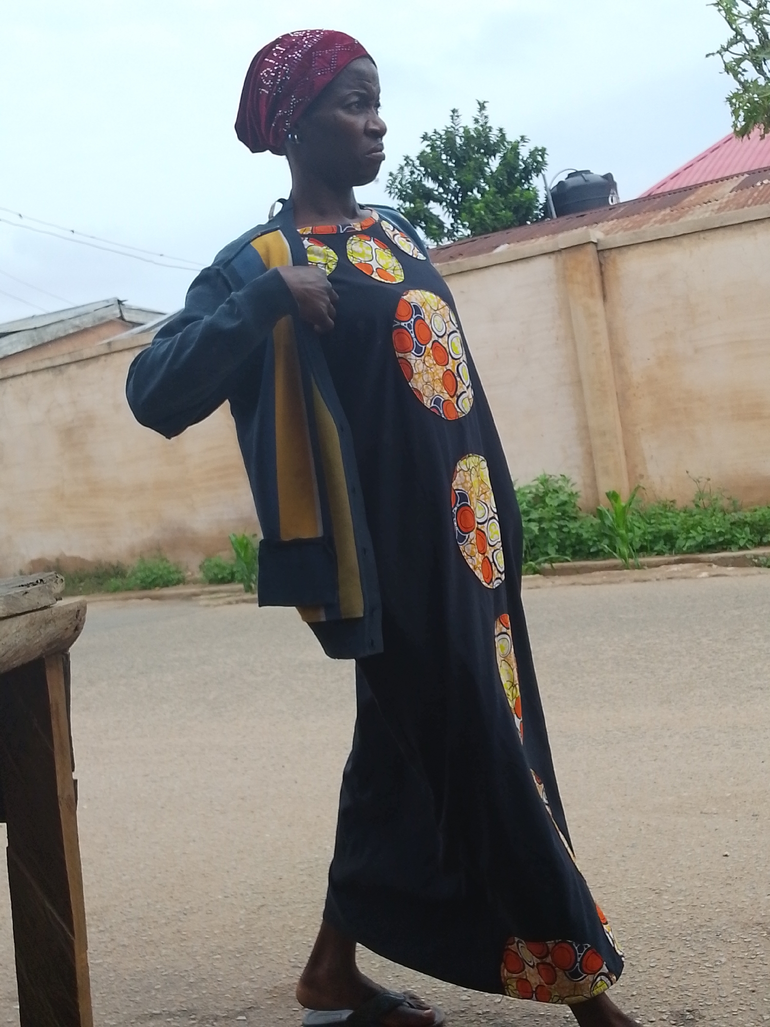 A middle-aged African woman is standing on a paved road in Jos, Nigeria. She is wearing a dark blue, ankle-length dress with circular patterns in shades of orange, yellow, and white. On her head, she has a dark red headscarf. She is holding a dark blue long-sleeved shirt with yellow trim on her left shoulder. She is looking towards the right side of the frame. The environment consists of a concrete wall along the side of the road with weeds and shrubs growing at its base. A water tank can be seen on top of a building with a corrugated red roof in the background. The image is taken during daylight hours under a cloudy sky.