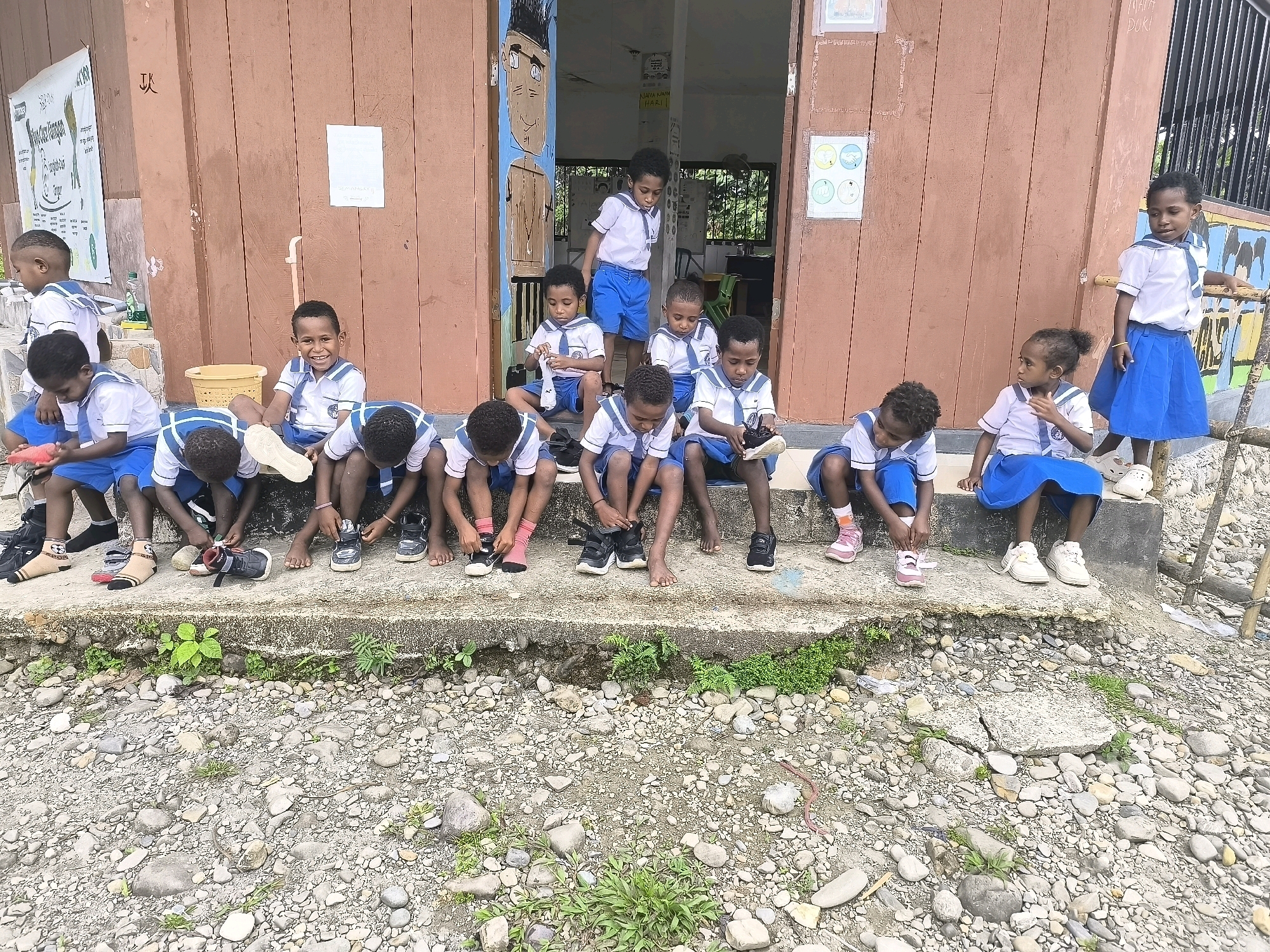 In Timika, Indonesia, a group of approximately ten young children, most wearing school uniforms consisting of white shirts with blue trim and blue shorts or skirts, are gathered on the steps of a building with a reddish-brown wooden exterior. Several children are sitting on the steps, some with their shoes off, while others are in the process of putting them on or taking them off. One child stands at the top of the steps, looking down. The building has an open doorway revealing a dark interior. A poster is visible on the wall to the left, and another smaller paper is taped to the wall to the right of the doorway. The ground in front of the steps is covered with small stones and pebbles, with some sparse green vegetation growing amongst them. A yellow bucket is visible on the left side of the steps. The overall scene appears to be an outdoor school environment.