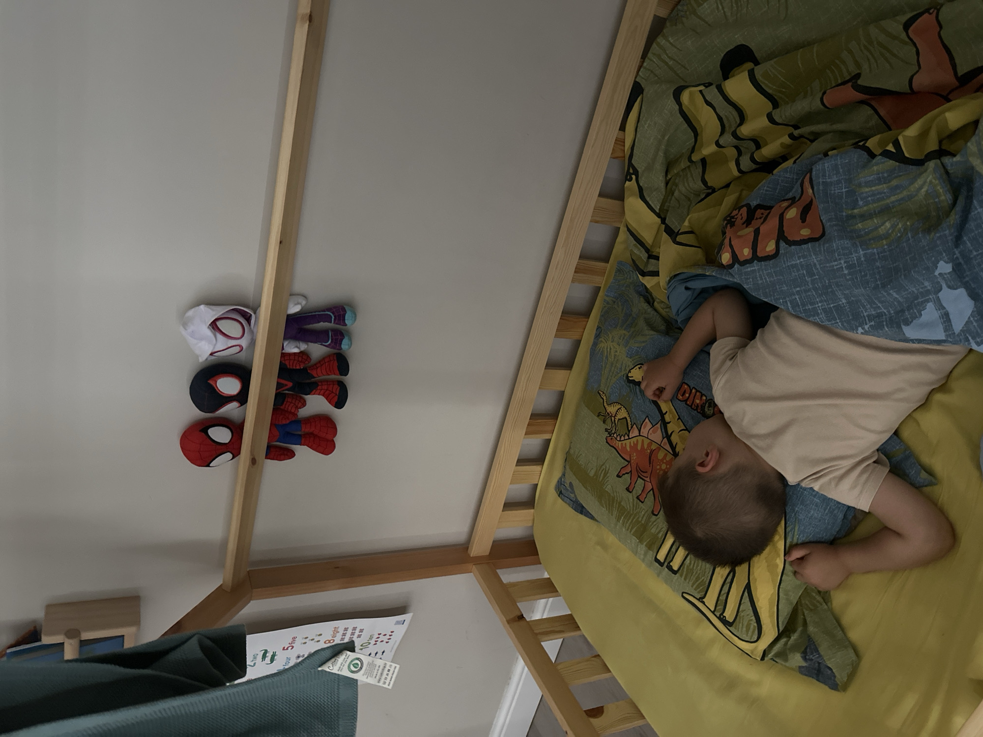 A young boy is sleeping in a wooden bed with a yellow sheet and a duvet featuring a dinosaur print. Three Spider-Man plush toys hang from a wooden railing above the bed. A teal garment is draped over the side of the bed. A poster with numbers and illustrations is attached to the wall. The scene appears to be in a bedroom.