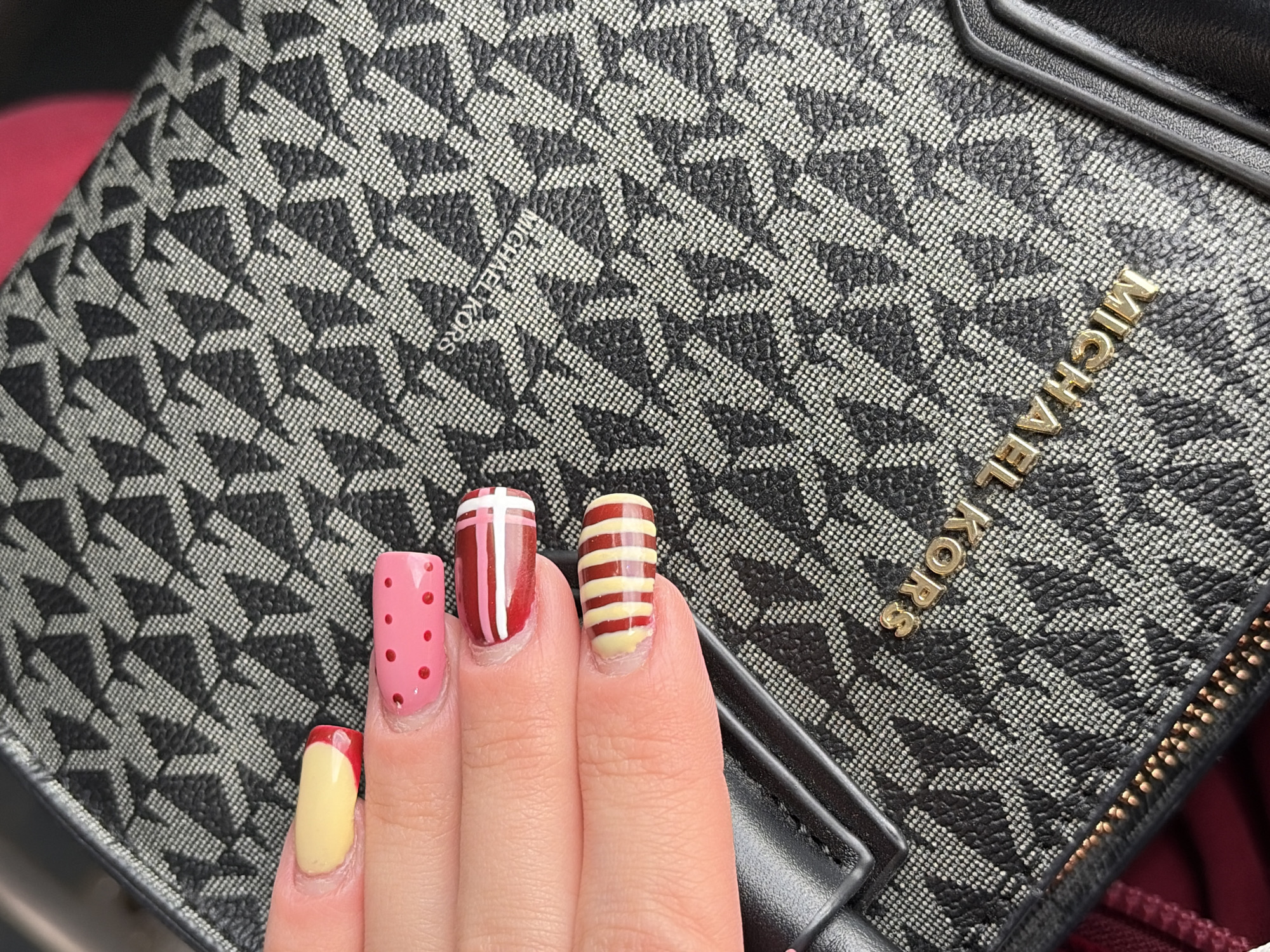 A close-up shot features a person's hand displaying a colorful and intricately painted manicure, resting on a black and grey patterned Michael Kors accessory, possibly a wallet or small bag. The image is set in a provided location context of Perm, Russia, though no specific visual cues within the image confirm this.
Four nails are visible, each with a unique design. The index fingernail is painted a light pink with scattered red polka dots. The middle fingernail has a deep red base adorned with white vertical and horizontal stripes, forming a grid or cross pattern. The ring fingernail is a light cream or yellow color with multiple horizontal red stripes. The pinky fingernail exhibits a light cream or yellow base with a bold red tip, resembling a French manicure.
The accessory serving as a backdrop is made of a textured material, featuring a repeating monogram pattern of "MK" initials in grey against a dark, possibly black, background. The brand name "MICHAEL KORS" is prominently displayed in raised gold-colored letters on the accessory's front surface and is also subtly integrated into the grey monogram pattern. A gold-toned zipper with visible teeth runs along the edge of the accessory. A blurred reddish-maroon area is visible in the background, suggesting an underlying surface or interior.