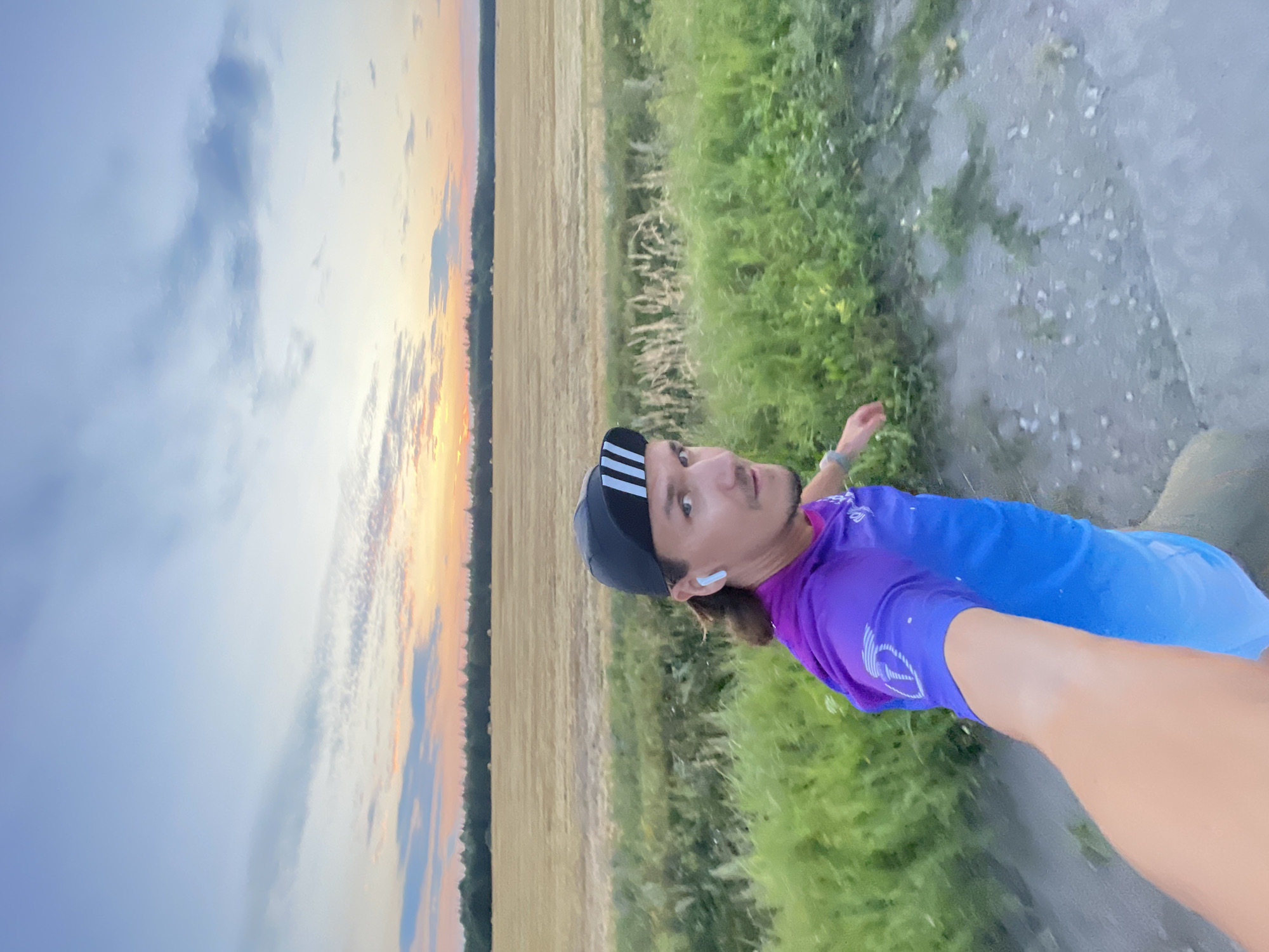 A man is taking a selfie with a long arm, as he runs along a dirt path. He is wearing a black cap with white stripes, a purple and blue ombre athletic shirt, and white wireless earbuds. His right arm is extended forward, capturing the image. Behind him, a field of harvested wheat stretches to a tree line, under a pale blue sky with hints of sunset colors. The ground is a mix of dirt and pebbles, with green grass and weeds growing along the side. The location is identified as a rural area outside of Unknown, Russia.