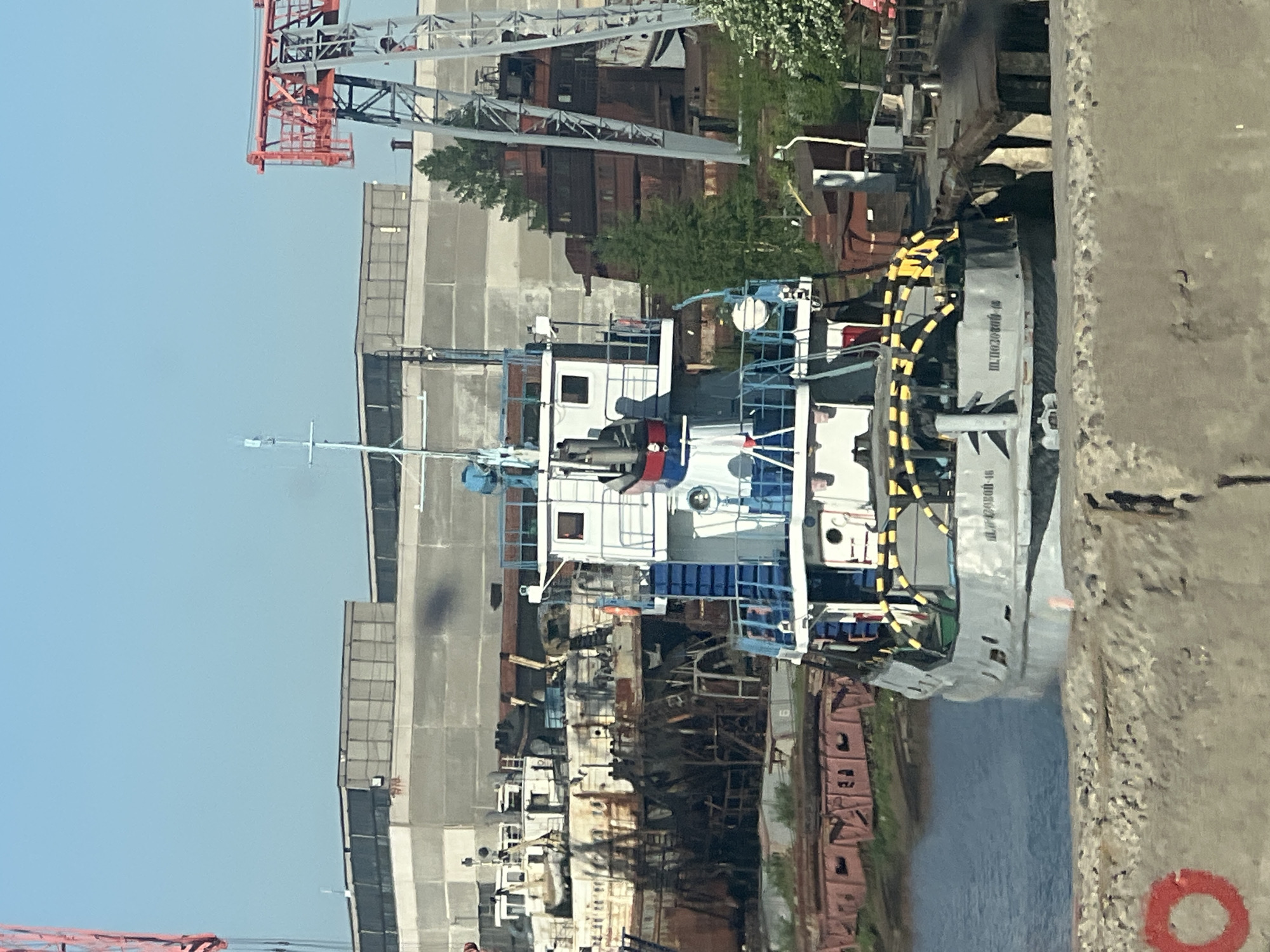 The image depicts a river port, specifically located in the city of Chaikovsky, Russia. A small, white tugboat is docked along a concrete pier. The hull of the tugboat is gray and it has yellow and black striped bumpers. The superstructure of the tugboat is white with blue railings. On the pier, a red circle is painted near the lower right corner of the image. Behind the tugboat, there are several large industrial buildings made of gray concrete. A red and gray crane is positioned near the buildings. The sky is clear and blue.