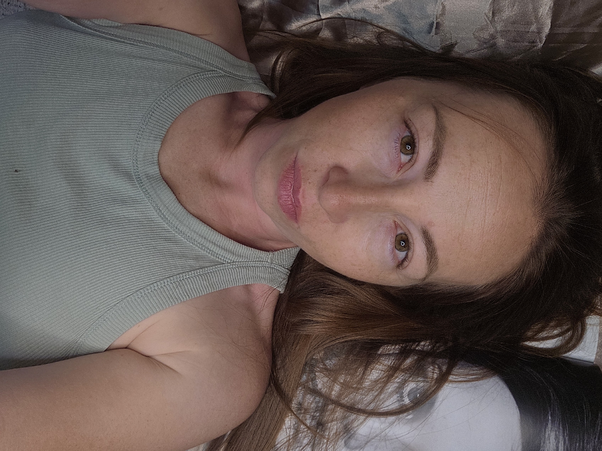 A close-up overhead shot shows a woman lying down and looking at the camera. Her head is tilted slightly and she has brown hair that is spread out around her. She is wearing a light green tank top. The background appears to be a bed with a textured gray comforter, and to the right of her head, there is a faint image of a face on a white surface. The image was taken in Chaikovsky, Russia.