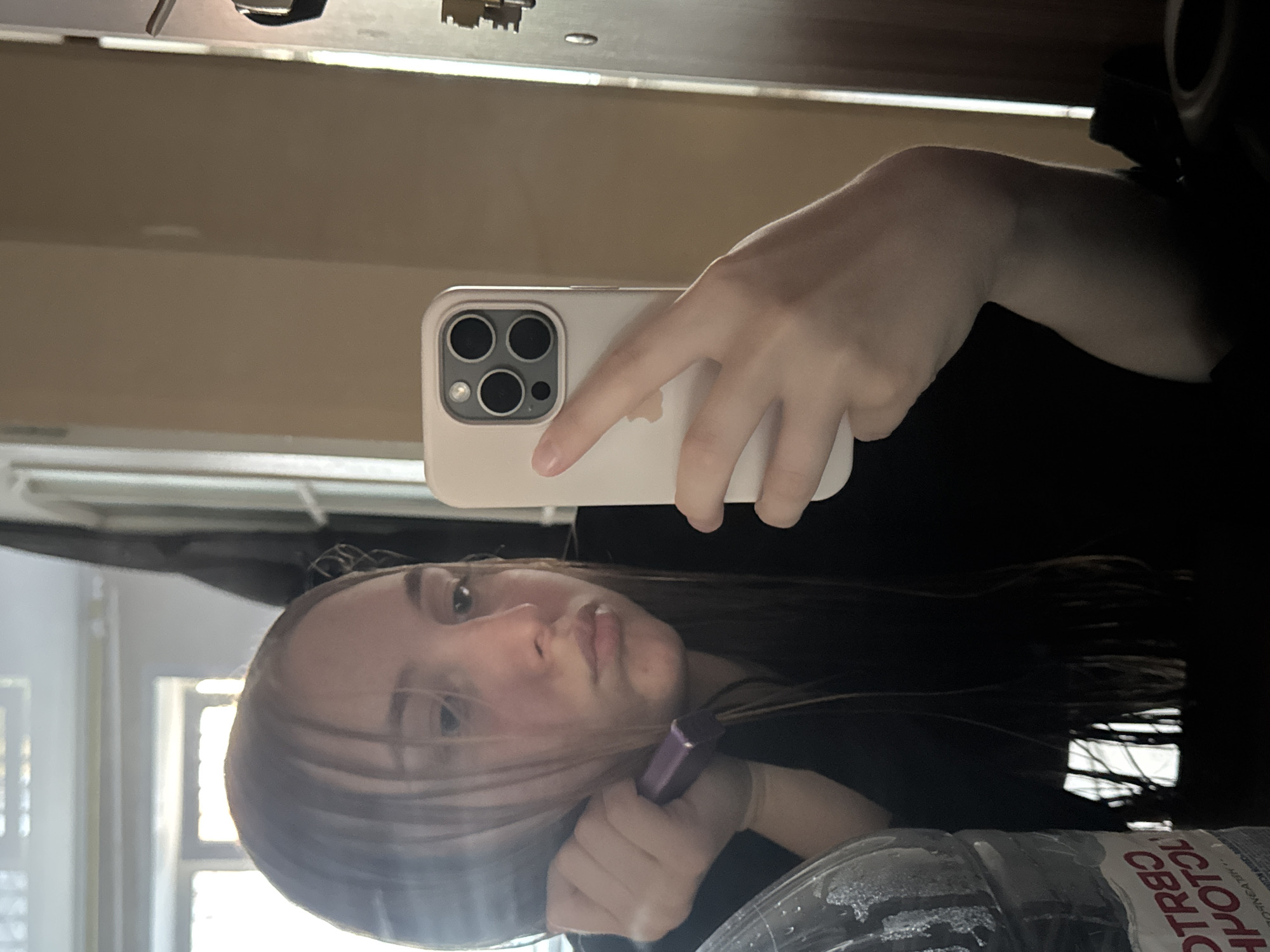 A young person, likely female, with long dark hair, captures a mirror selfie indoors. They hold a light-colored iPhone, identifiable by its camera module and a faint Apple logo on the case, in their right hand. In their left hand, they hold a small purple cylindrical object, possibly a lipstick or hair accessory, near their hair. The person's expression is neutral with slightly pursed lips.
The scene appears to be an indoor setting, likely a room, with natural light streaming through a window visible in the reflection behind the subject. The overall lighting suggests it is daytime. In the foreground, a partially frosted clear plastic water bottle is visible, featuring distinct Cyrillic text. The word "СВЯТОЙ" (Svyatoy - Holy) is clearly legible on the bottle's label, indicative of a Russian brand like "Святой Источник" (Holy Spring water). This detail, combined with the context of Perm, Russia, reinforces the setting.