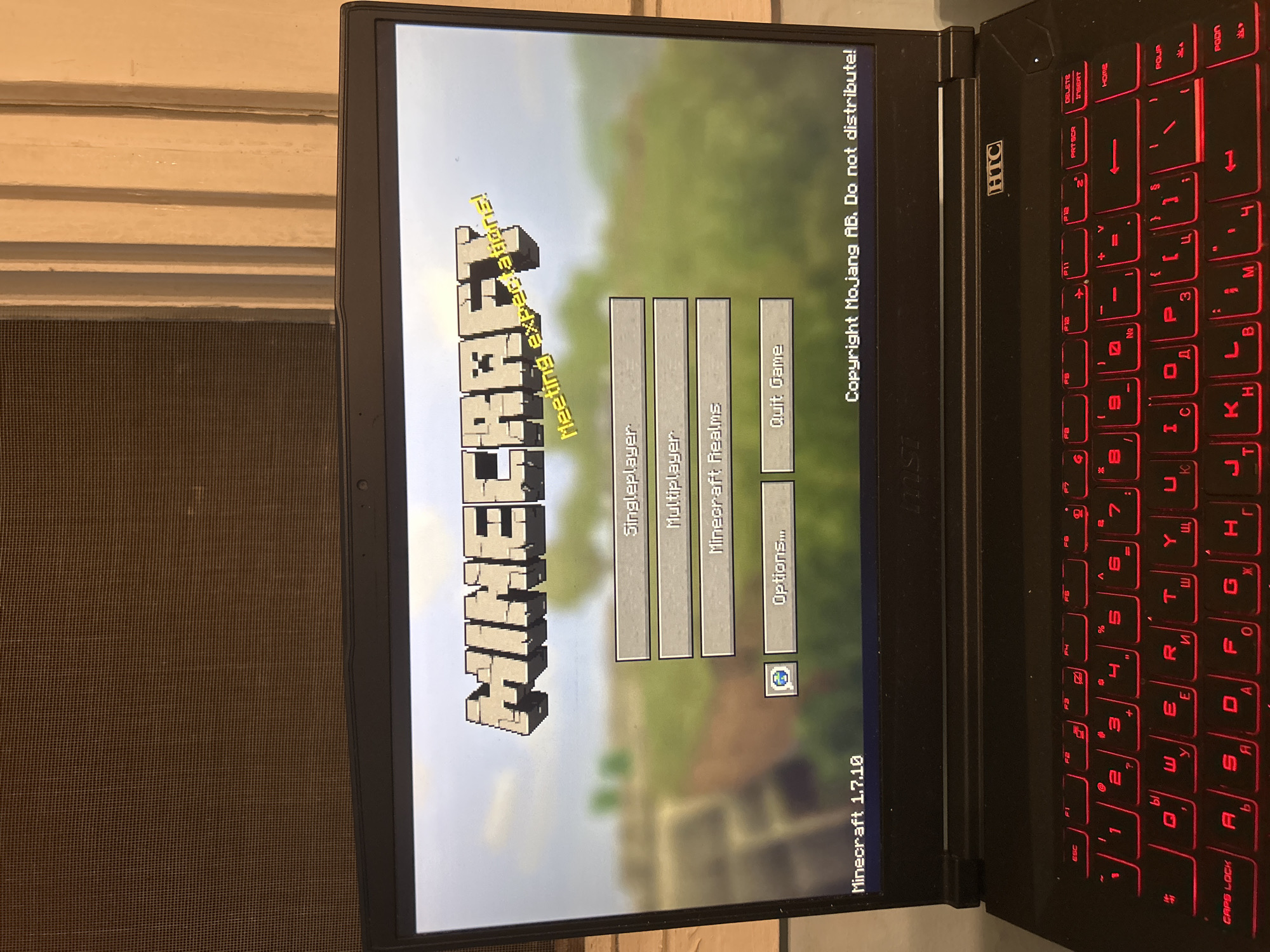 The image shows a laptop screen displaying the main menu of the video game Minecraft version 1.7.10. The menu options visible are Singleplayer, Multiplayer, Minecraft Realms, Options, and Quit Game. The game logo is prominently displayed at the top of the screen with the tagline "Meeting expectations!". The screen also displays the copyright information for Mojang AB. The laptop itself is black and the keyboard has red backlighting. The laptop appears to be on a surface in front of what looks like a window screen and a light-colored wall. The location in the city of Unknown, Unknown, is not discernible from the image.