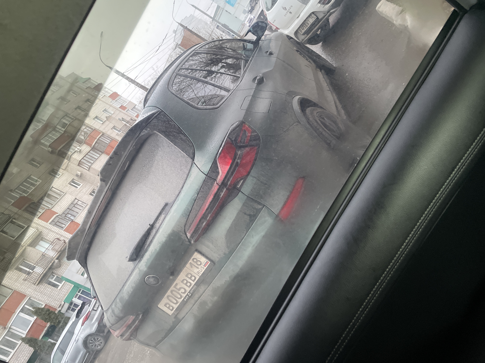The image, taken from inside a vehicle, shows a gray car parked on a street. The car's license plate reads "B005 BB 18 RUS," identifying it as Russian. Buildings are visible across the street. The location is Izhevsk, Russia.