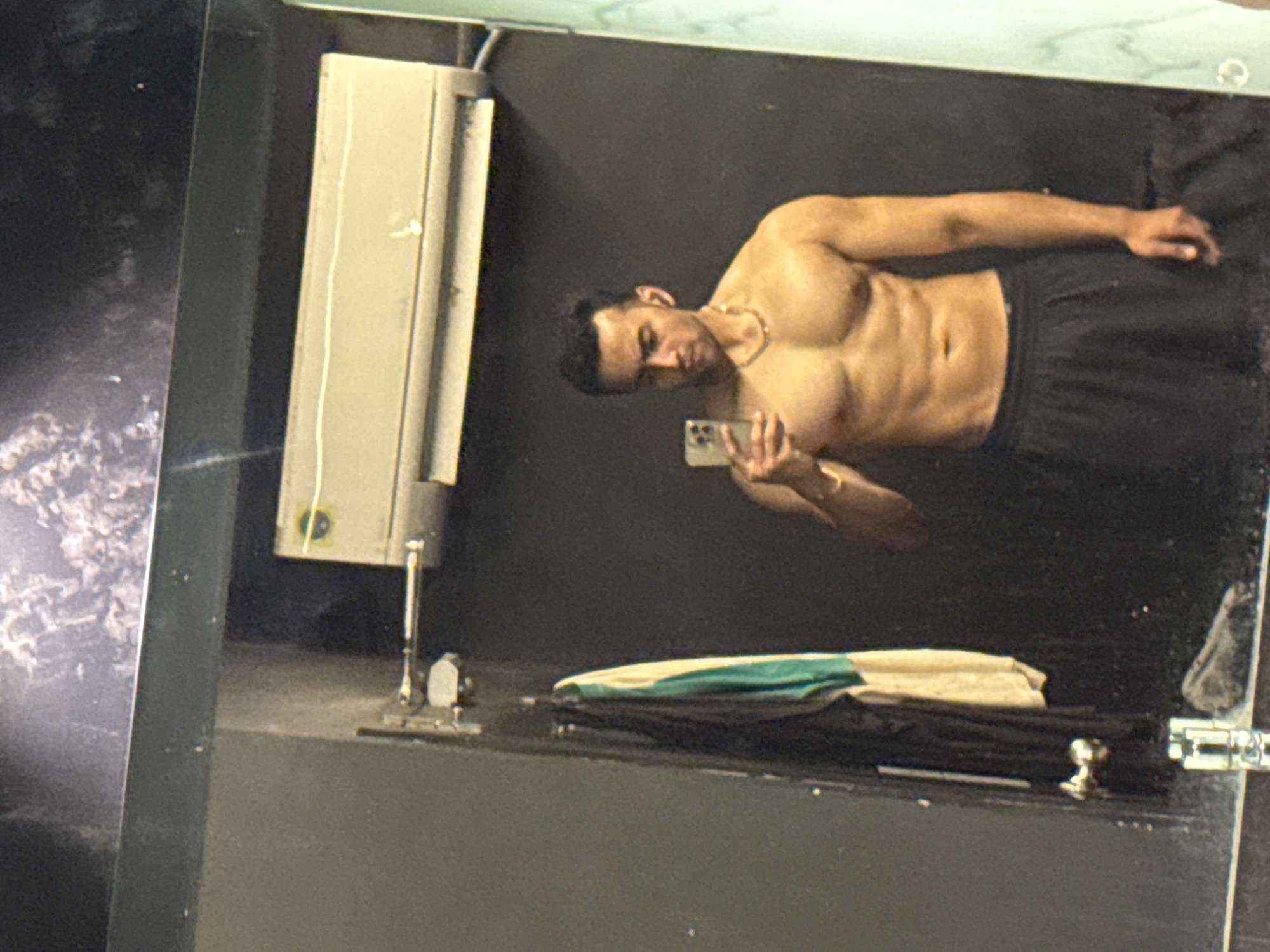 The image is a selfie of a shirtless man in a mirror. He is holding a phone in his left hand, presumably taking the picture. He is wearing a gold necklace. The background is dark, with a light fixture on the left side of the frame. There's also clothing laying on a surface in the foreground. There are no visible elements indicating the location is Kolkata, India.