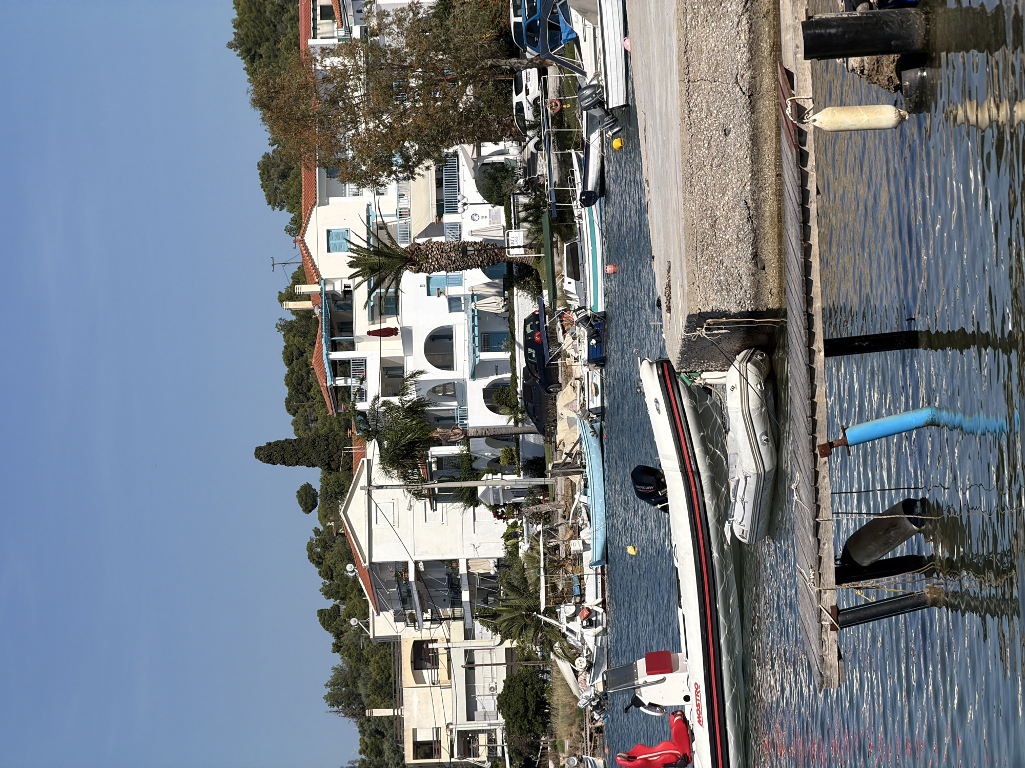 This image captures a vibrant, sunny waterfront scene in a coastal town, consistent with the location context of Poros, Greece. The perspective looks across a body of water towards a row of buildings and a dock.
On the right side of the image, calm, clear blue water shows gentle ripples, reflecting the bright sky. A concrete walkway and a wooden pier stretch along the water's edge. Numerous boats are moored here, varying in size and type. In the foreground, a white inflatable boat with a red stripe, bearing the partial text "MOST RO", is prominent, equipped with a "MERCURY" outboard motor. Next to it, another smaller inflatable boat is docked. Further along the pier, other boats are visible, one featuring a "YAMAHA" outboard motor. Various buoys in white, yellow, blue, and black are floating in the water or secured to the pier.
On the left, a series of multi-story white buildings with traditional red-tiled roofs line the waterfront. These structures feature balconies, some with light blue window shutters or railings, and appear to be residential or hospitality accommodations. Lush vegetation, including several tall palm trees and other green trees, is interspersed among and behind the buildings, adding to the Mediterranean aesthetic. Cars are parked along what appears to be a road or access path fronting the buildings.
The time of day is clearly daytime, likely midday or early afternoon, given the bright, direct sunlight and clear blue sky. No people are visible in the scene, contributing to a sense of peacefulness and tranquility. The setting suggests a picturesque holiday destination or a quiet harbor town.