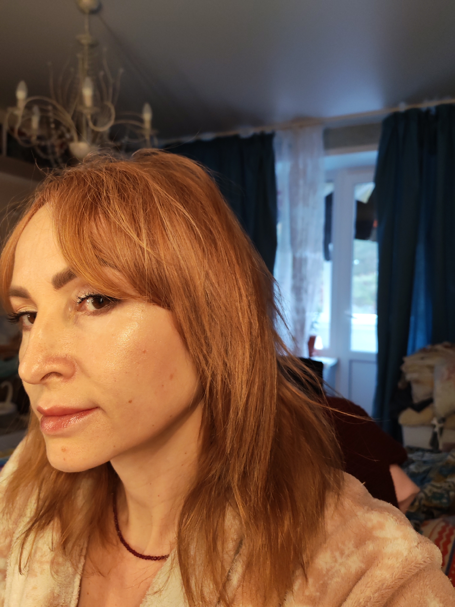 A woman with reddish-blonde hair, wearing a plush robe and a red beaded necklace, is posing for a selfie. Her face is turned slightly towards the left, showing her profile. There is soft, natural lighting on her face, accentuating her features. The background includes a chandelier, a window with sheer and blue curtains, and a cluttered area with clothing or bedding. The location may be in Chaikovsky, Russia, but there's no way to corroborate that from the given image.