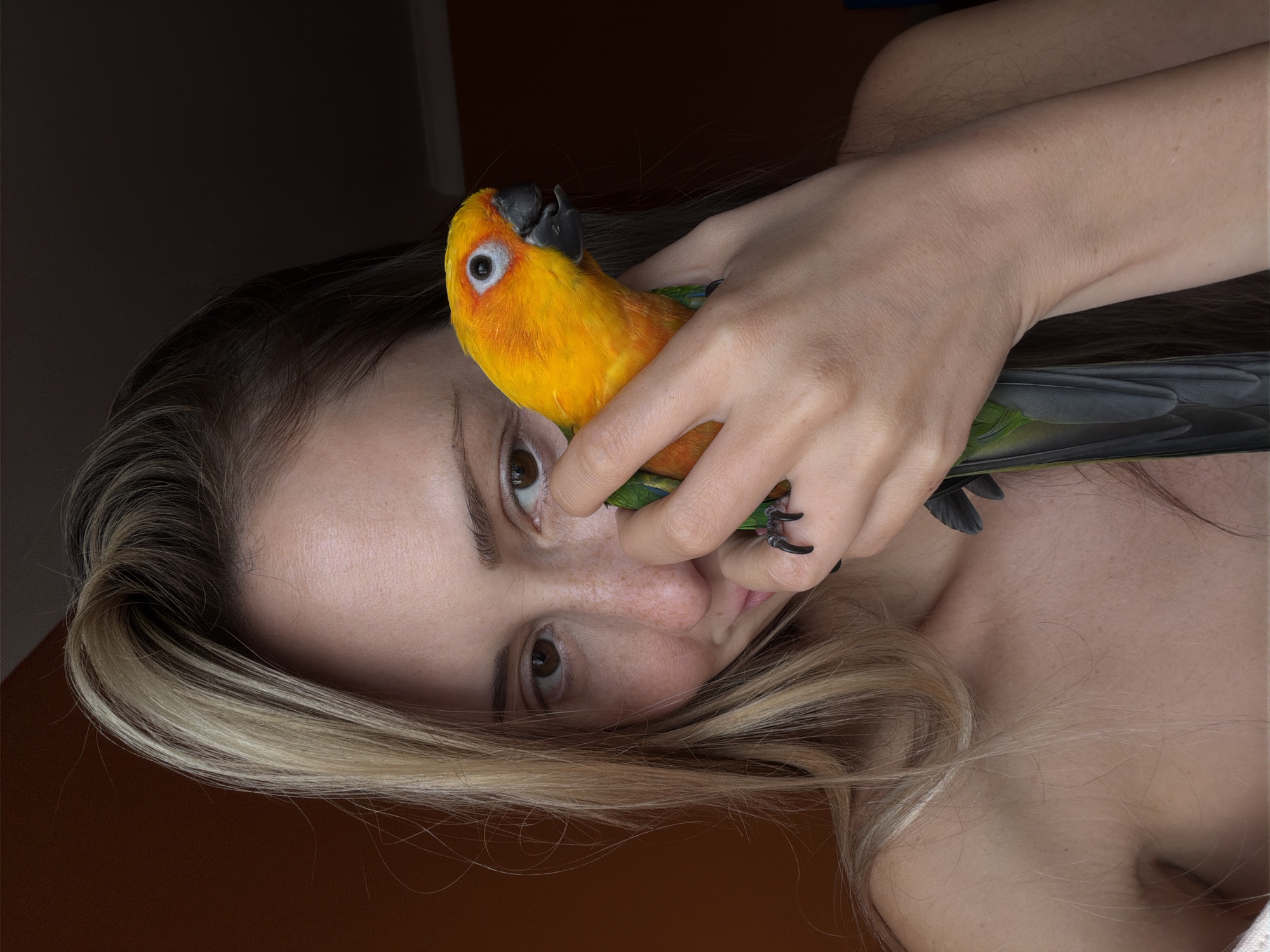 A close-up, head-and-shoulders portrait shows a young woman with blonde hair, some strands of which fall across her face. She is holding a bright orange and green parrot in her hands, and the bird is partially obscuring her face, with its head peeking out near her eye. The woman is looking downwards and slightly to her left, with a faint smile. Her skin is visible, and she has freckles on her nose and cheeks. The background is out of focus and appears to be an indoor setting with an orange-toned wall and a darker area. The image's lighting suggests it was taken indoors with natural light from the right. The context of Khimki, Russia, is not visually evident in the provided image.