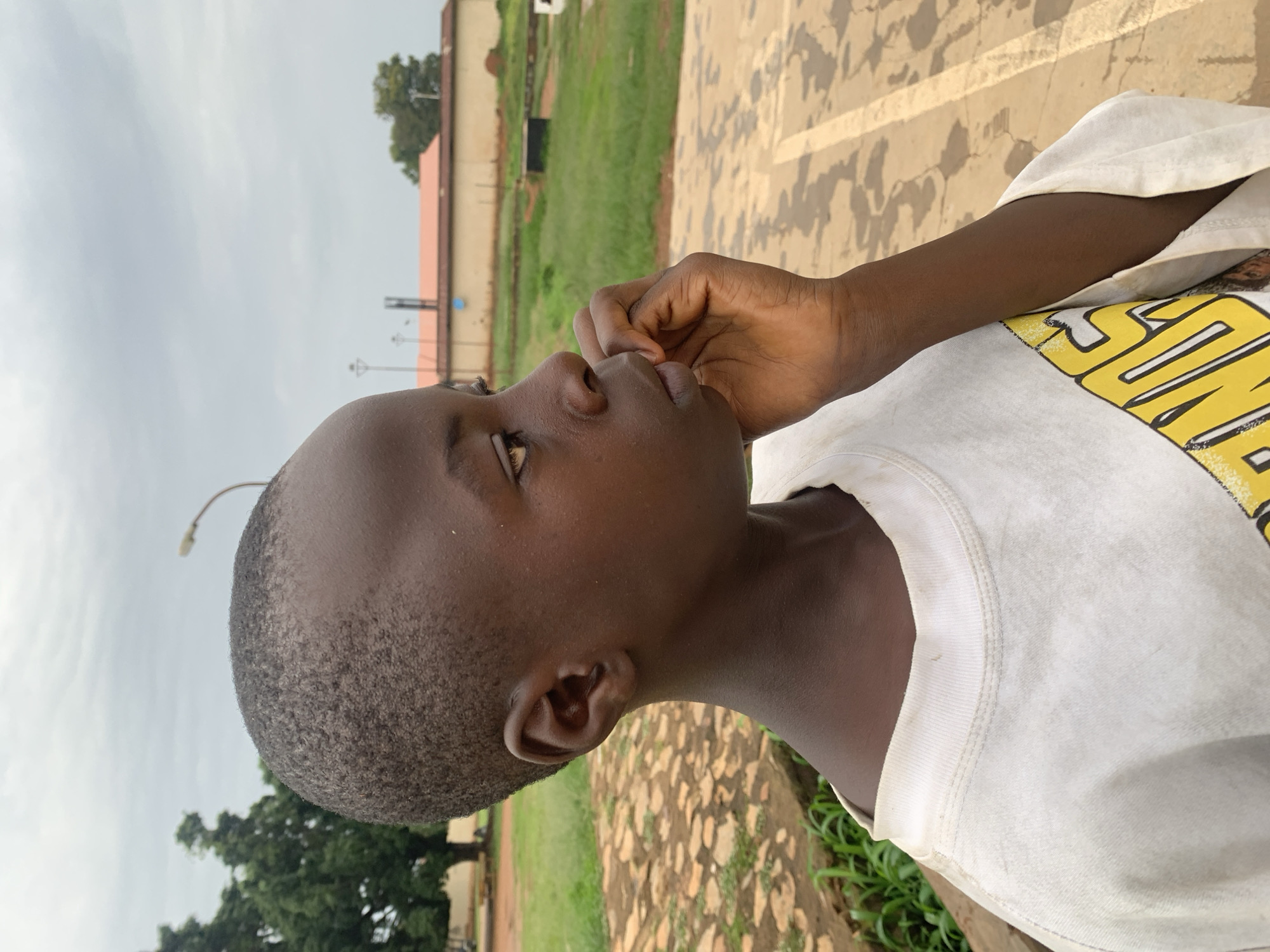 A close-up, profile view captures a young boy with dark skin and short, textured hair looking to his right. He wears a white t-shirt with a graphic design visible on the lower right side. His right hand is raised to his mouth, with his index finger touching his lips. The background suggests an outdoor, possibly urban or suburban, environment in Gura Top, Nigeria, with a paved area, grassy patches, trees, and a building in the distance under a cloudy sky.